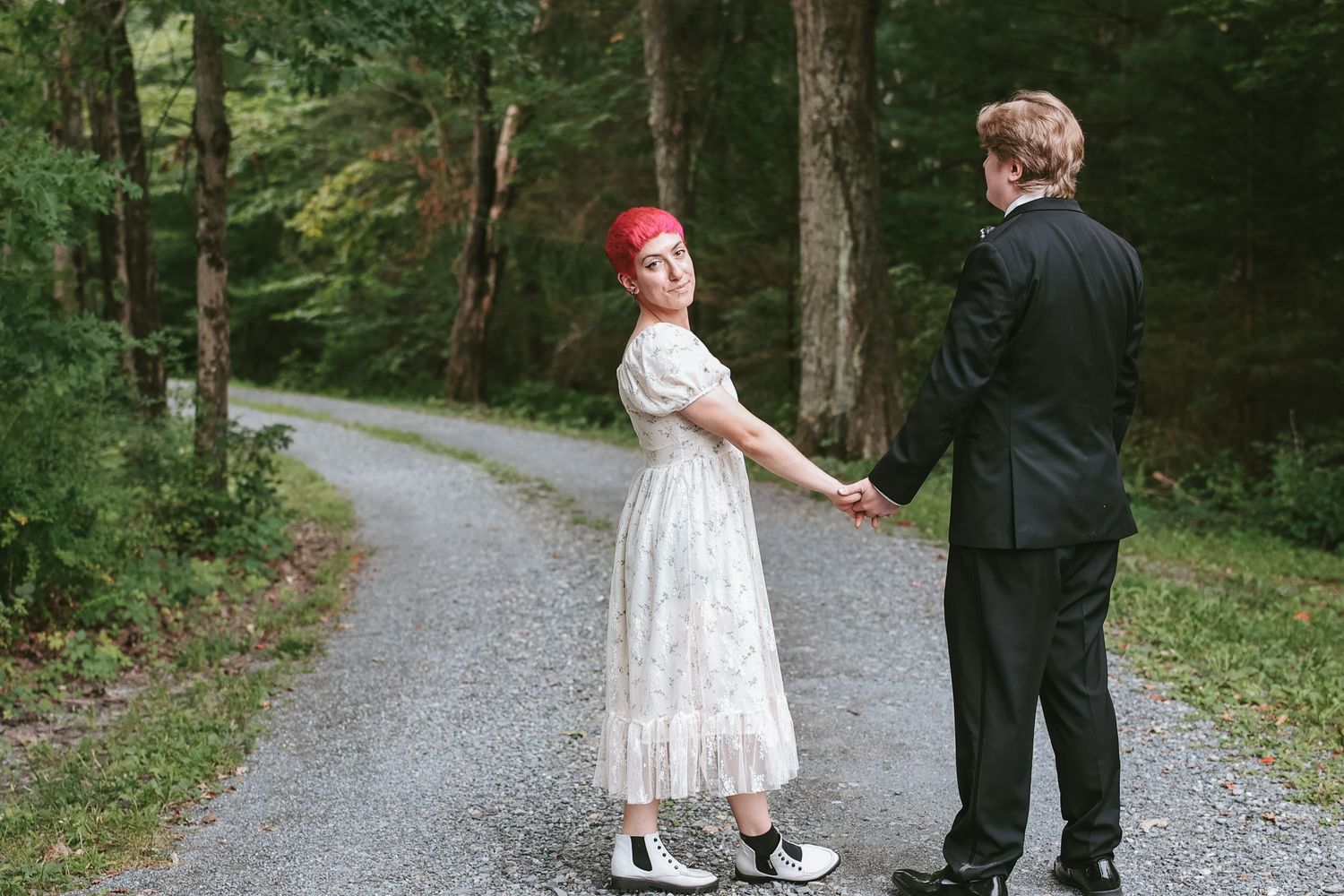 A couple in formal attire holding hands on a winding forest path, with the person in white wearing a red headpiece.