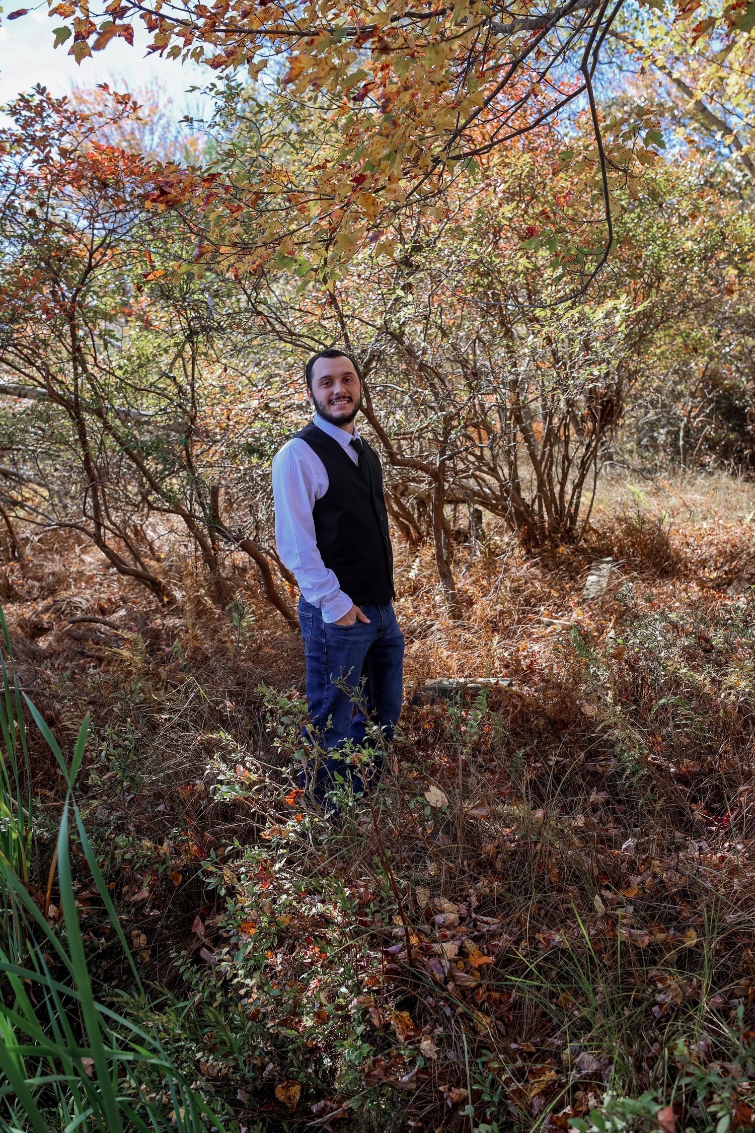 A person stands in autumn foliage wearing a vest and white shirt among fall colored trees and brush.