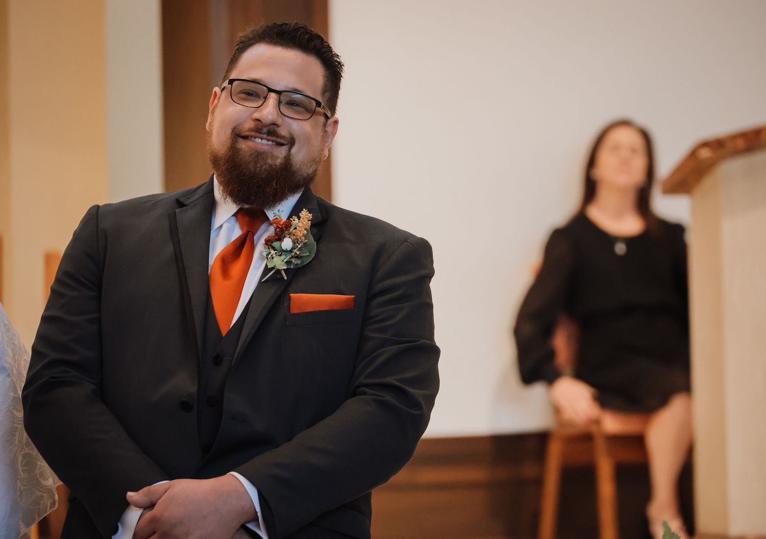 A person in a dark suit and orange tie stands in a formal setting during what appears to be a wedding ceremony.