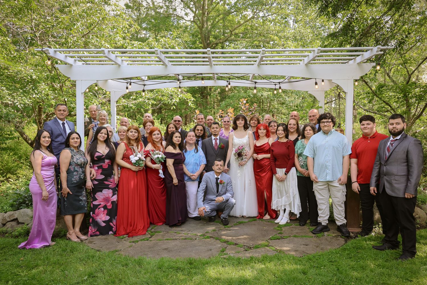 Large wedding party poses outdoors under white pergola for group photos in garden setting.