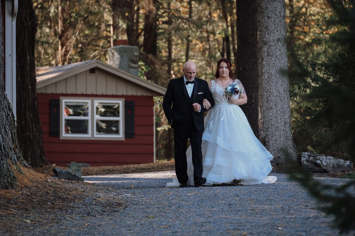 Wedding party walks along a wooded path outside a rustic red cabin during an outdoor ceremony.