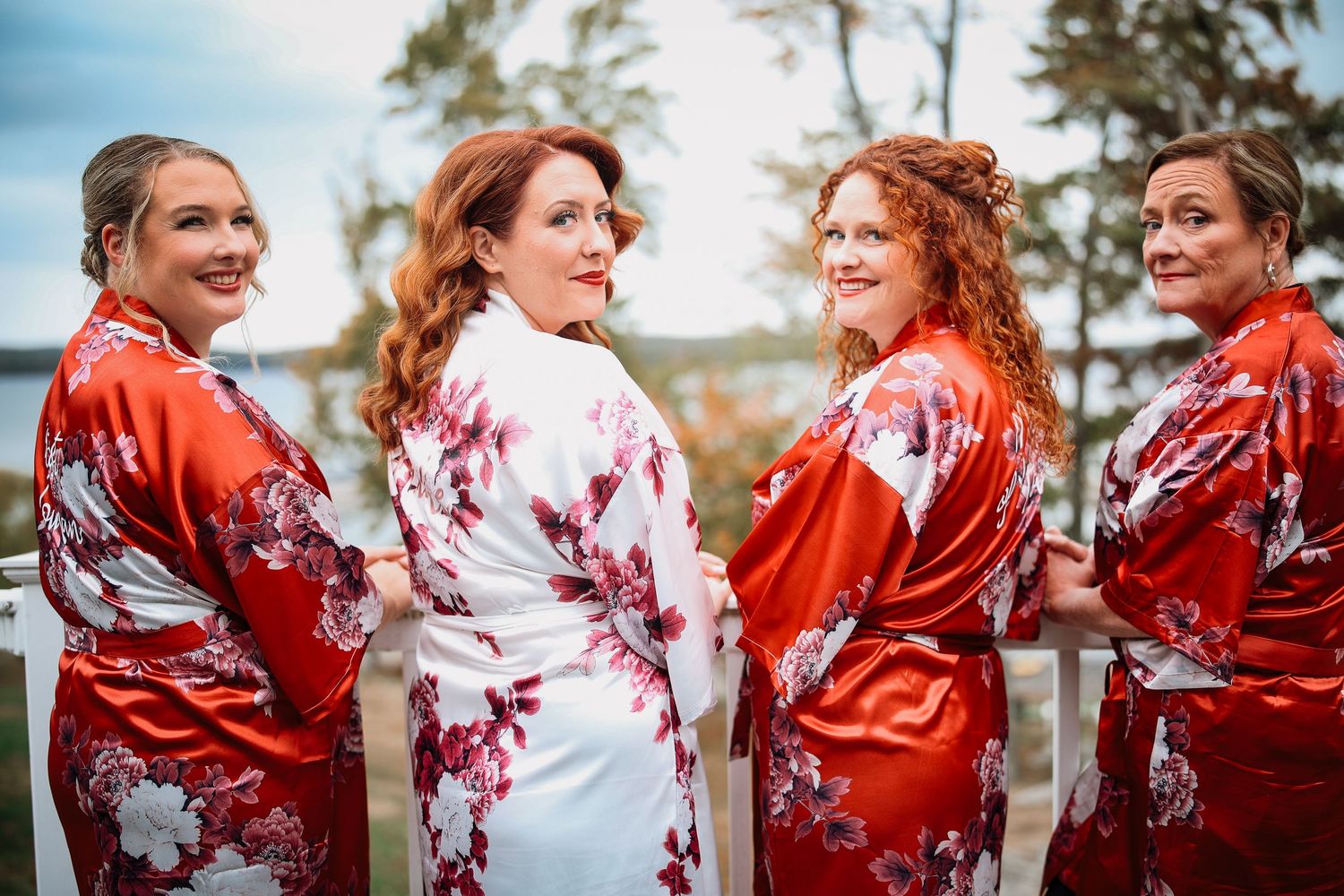 A group of bridesmaids in matching red and white floral robes laugh together during wedding preparations.