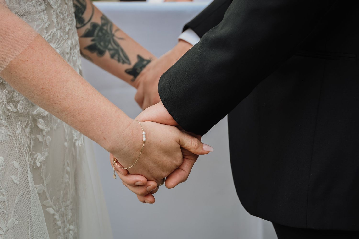 Close-up of newlyweds holding hands at wedding ceremony with visible tattoo on bride's arm.