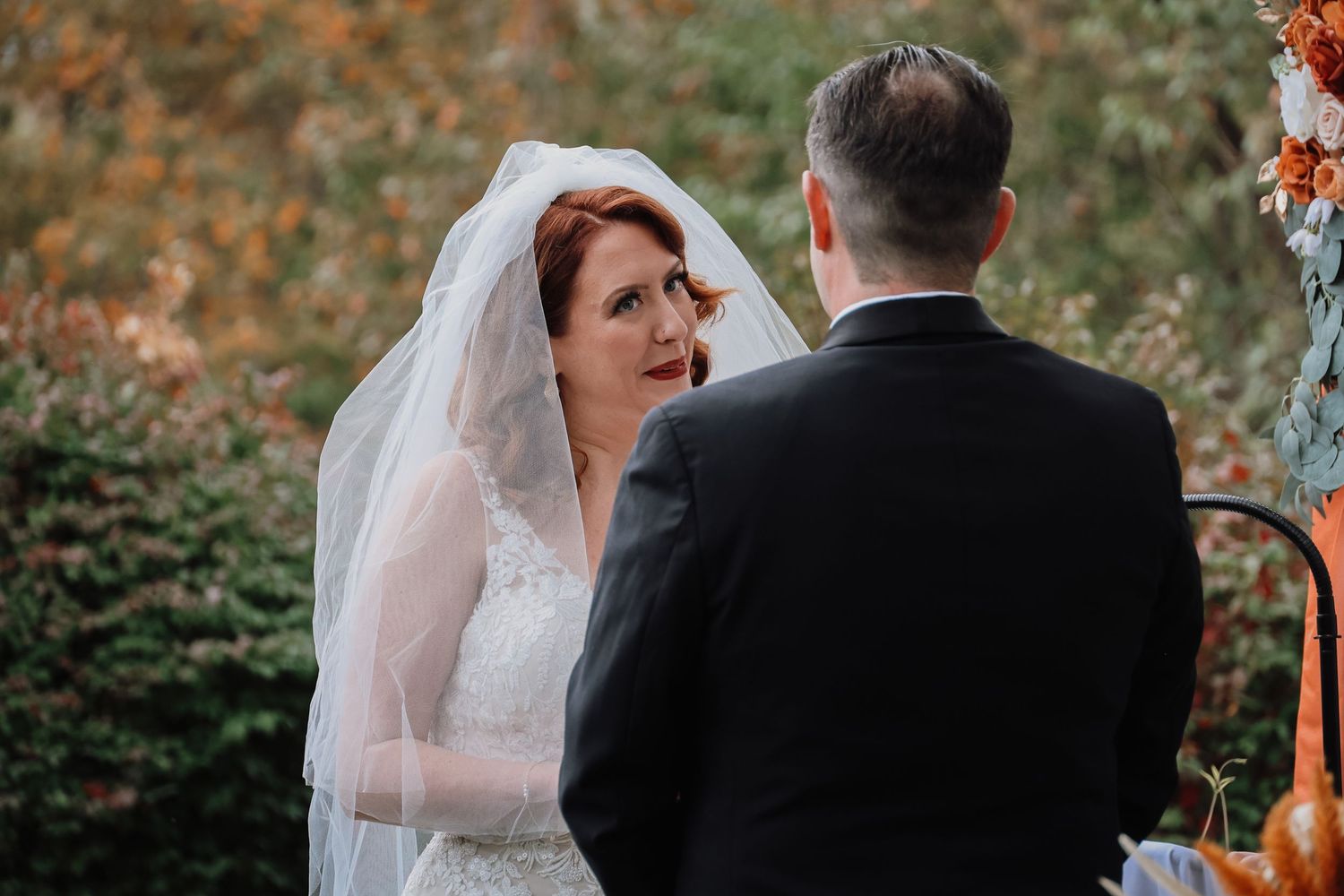 A bride in a white lace dress and veil looks lovingly at her groom during an autumn outdoor wedding ceremony.