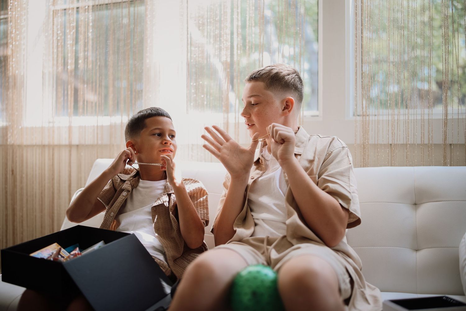 Two people sit on a white couch having an animated conversation in a sunlit room with sheer curtains.