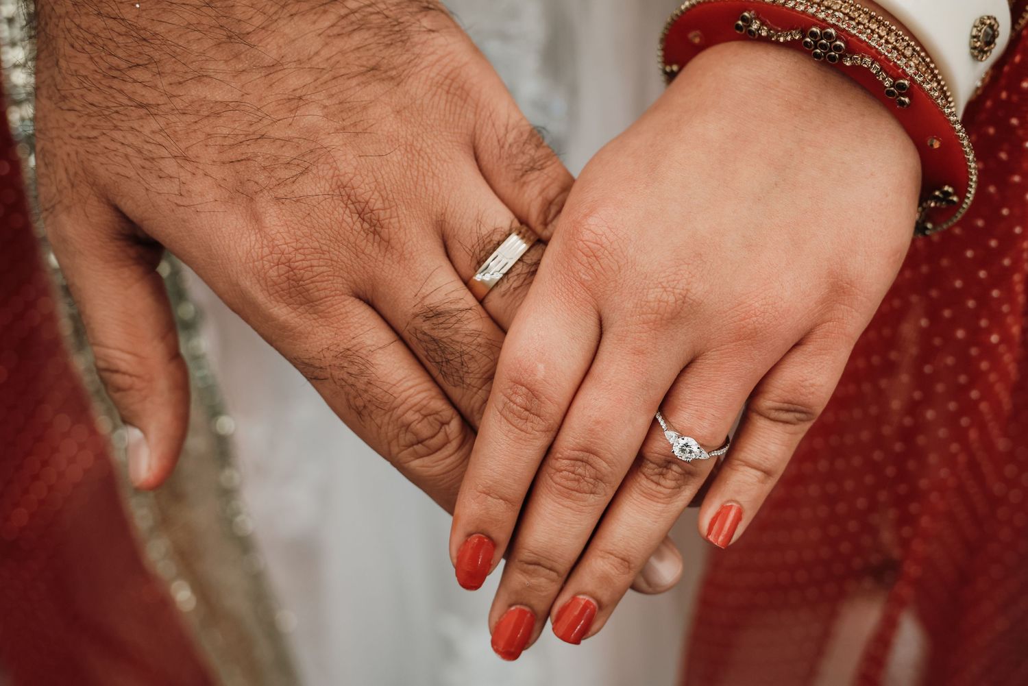 Close-up of hands wearing wedding rings with red nail polish and traditional Indian bridal jewelry.