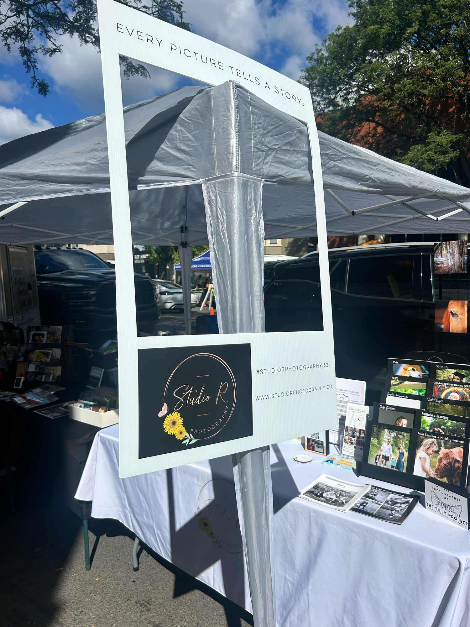 White popup tent displays with photo merchandise set up at an outdoor market or event space under sunny skies.