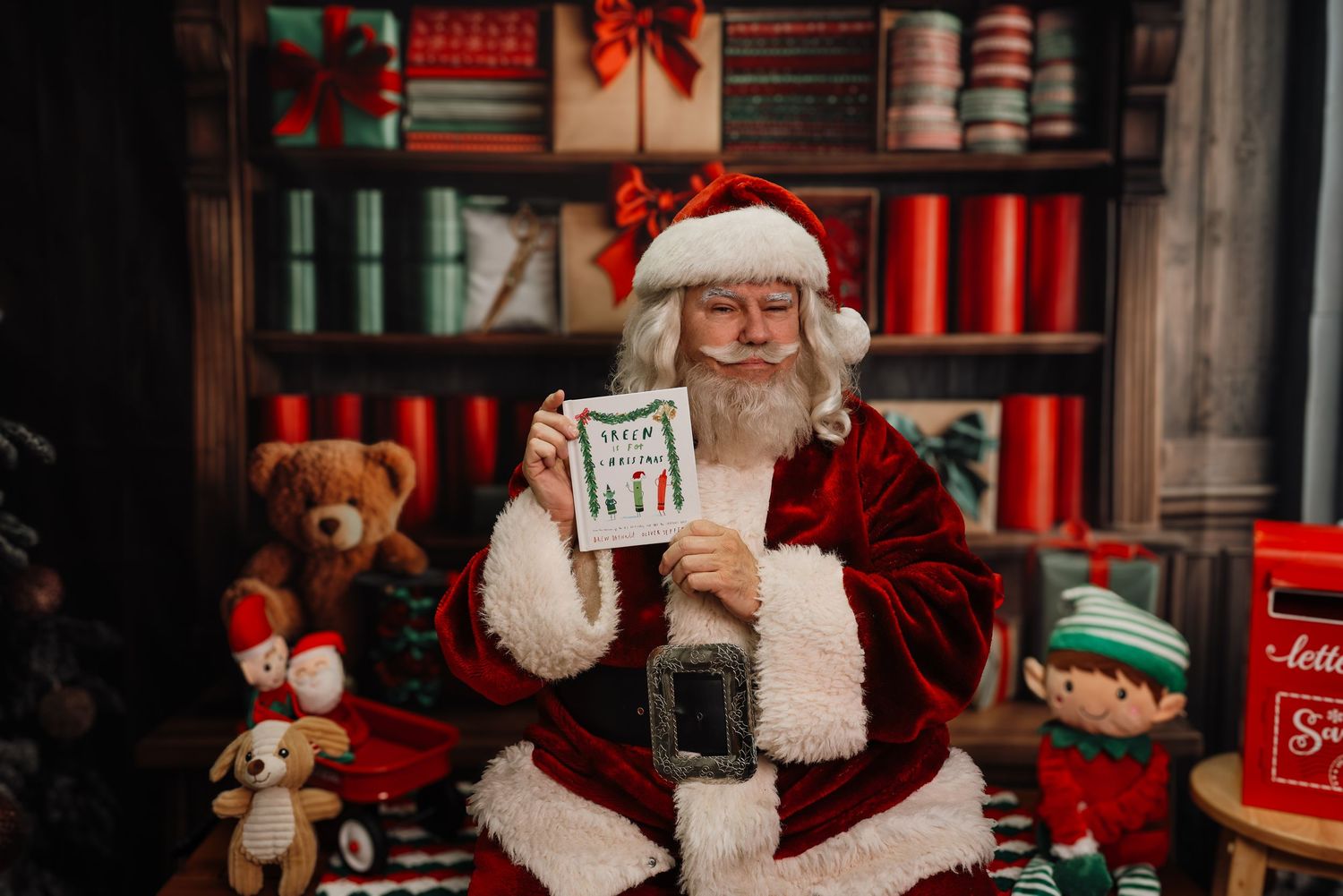 Santa and an elf helper sit at a desk in a cozy workshop filled with Christmas decorations and toys.
