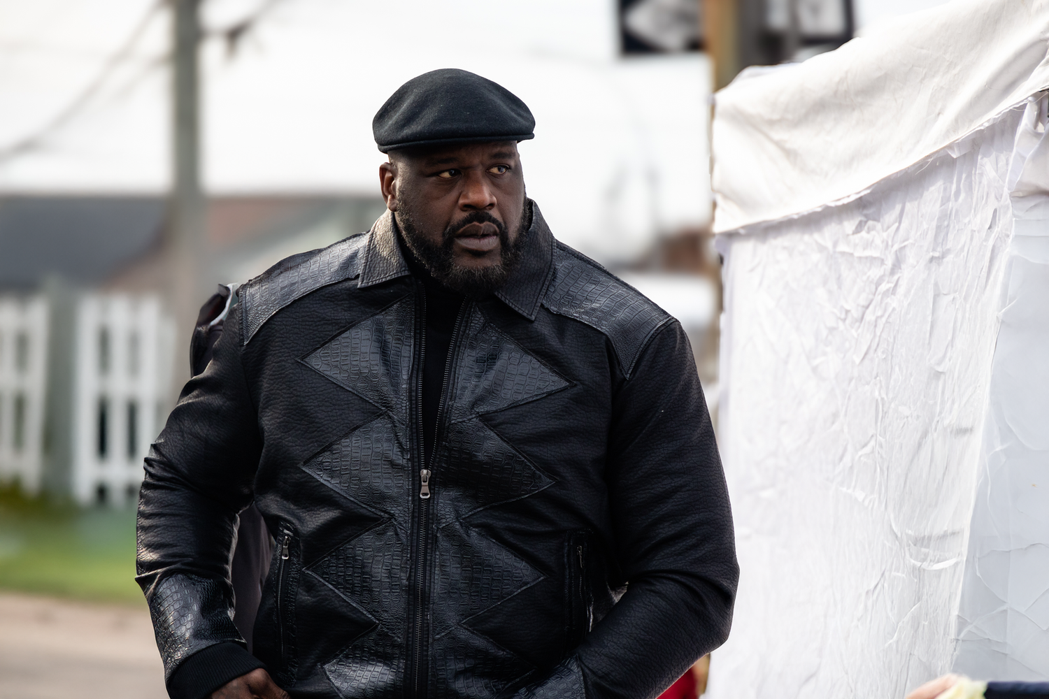 A person wearing a black winter coat and flat cap stands next to a white tent on an overcast day.