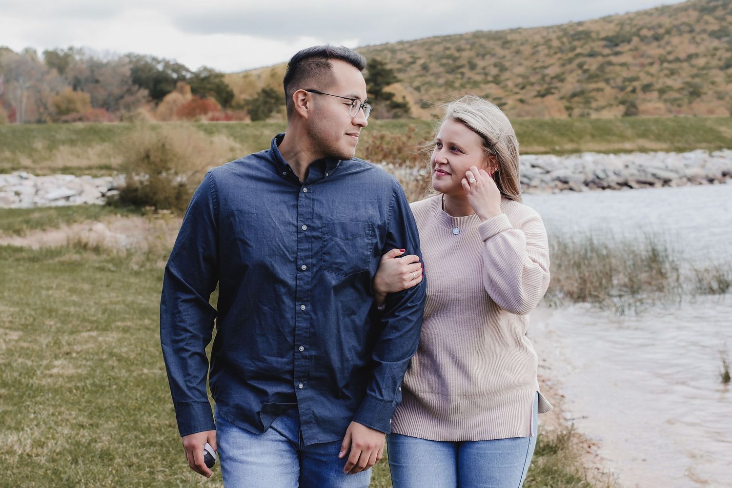 A couple shares a romantic moment while walking along a scenic lakeside path on a cloudy autumn day.
