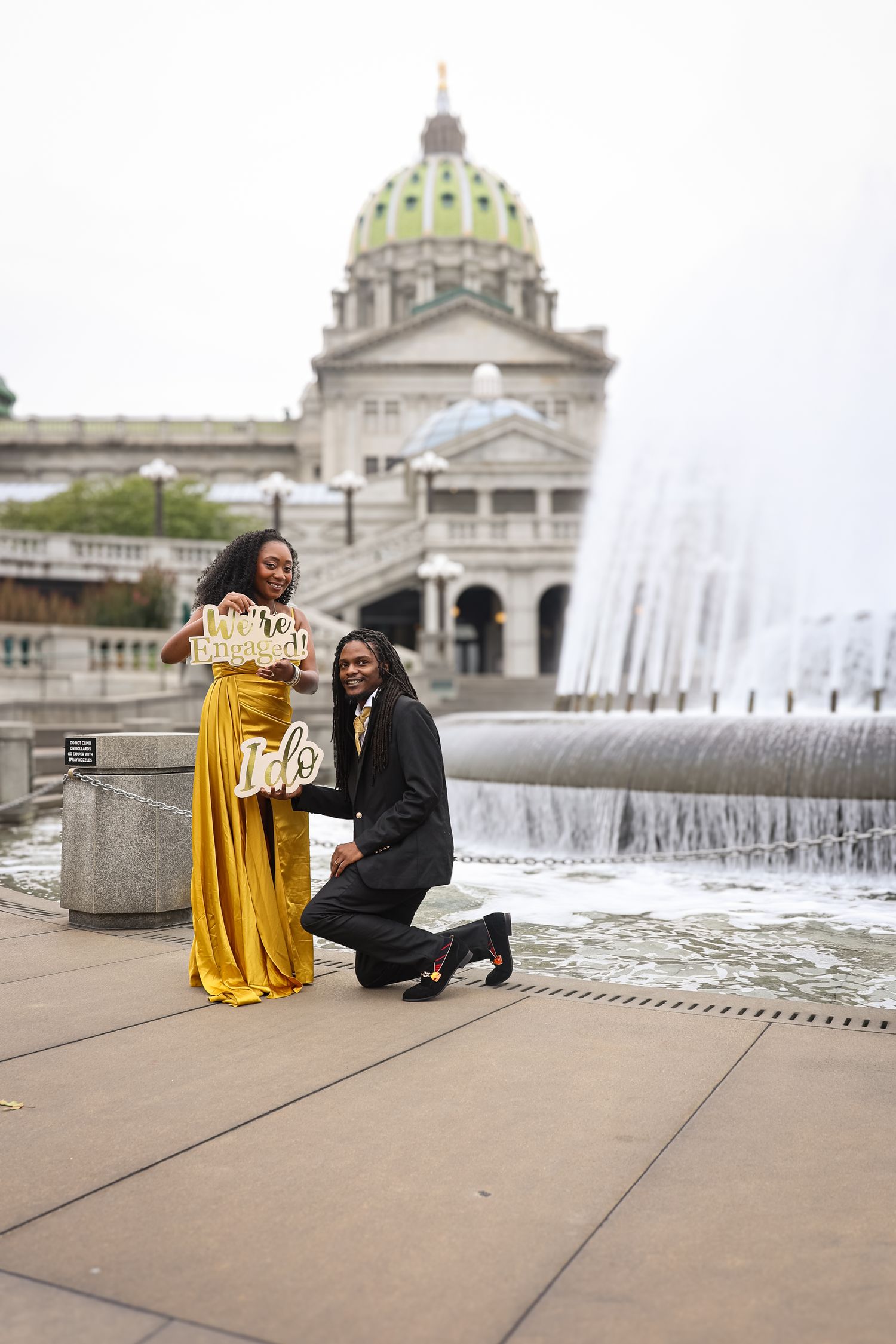 A romantic proposal moment in front of the Pennsylvania State Capitol building with a fountain in the background.
