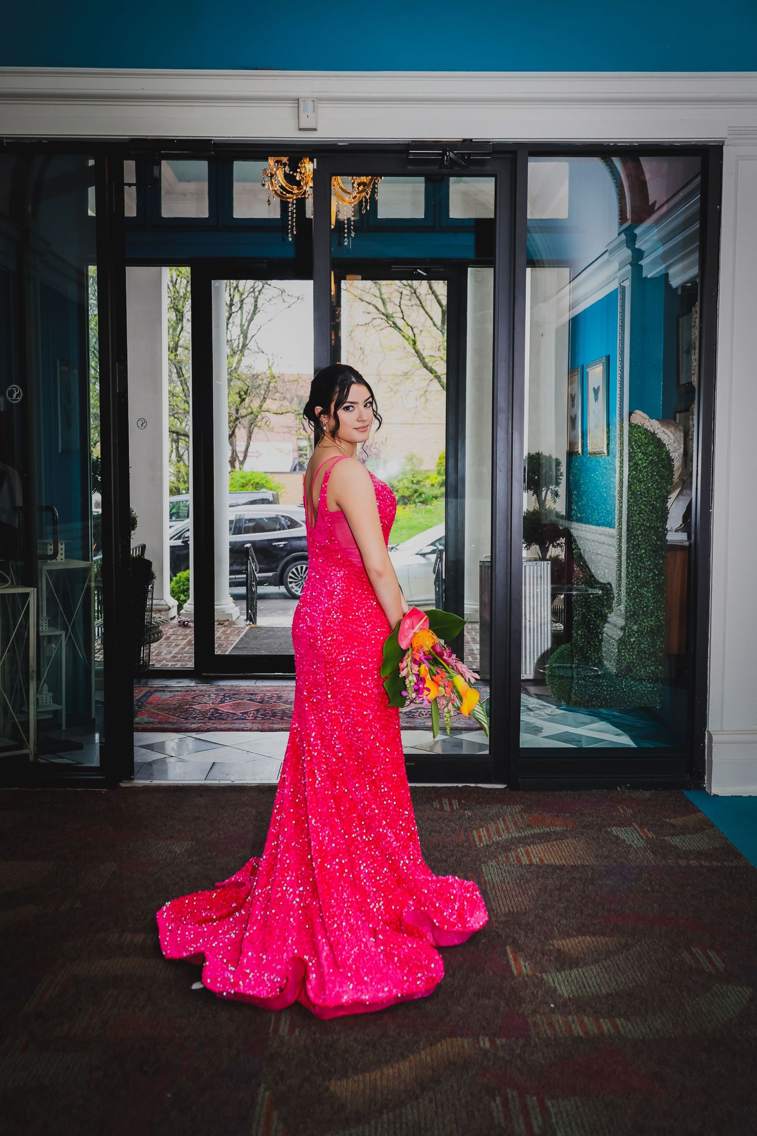 Woman in a stunning hot pink sequin mermaid gown with train holds flowers, posing at elegant venue entrance.