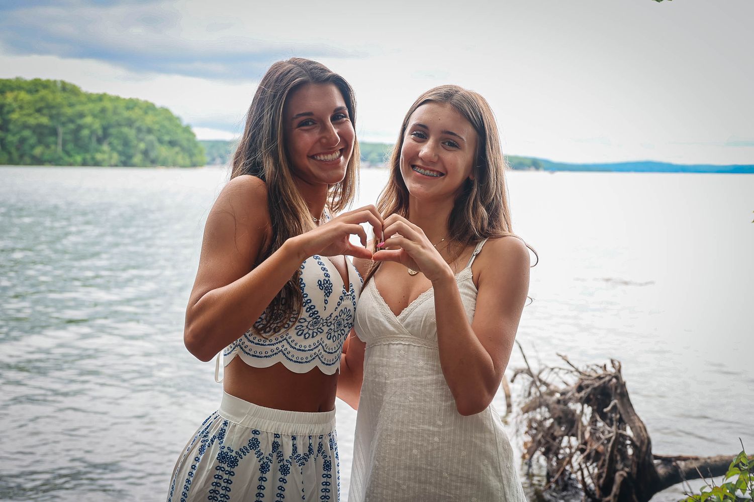 Two young women in white outfits make a heart shape with their hands by a scenic lakeside with lush green trees.