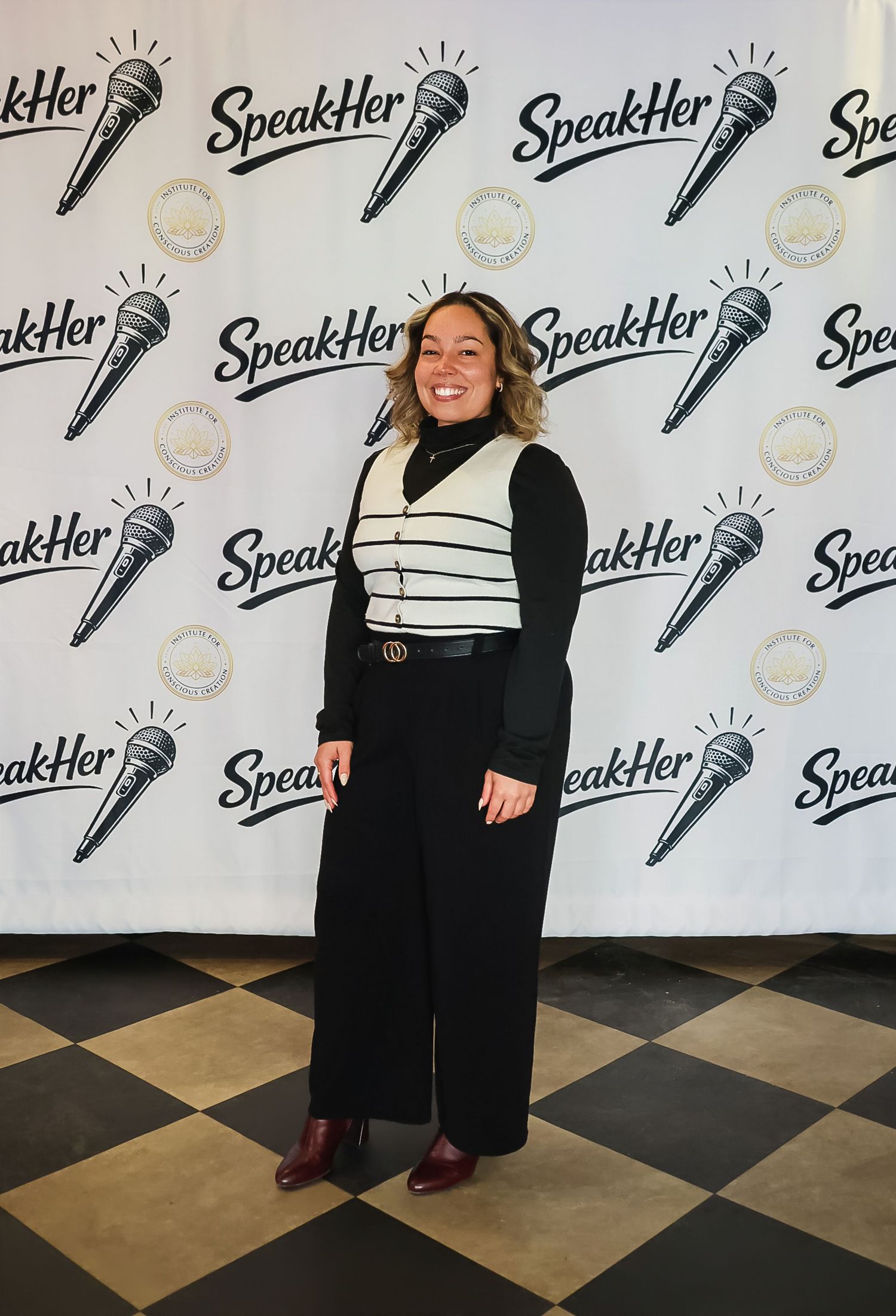 Woman smiling in black outfit with striped vest posing at SpeakHer event backdrop with microphone logos.