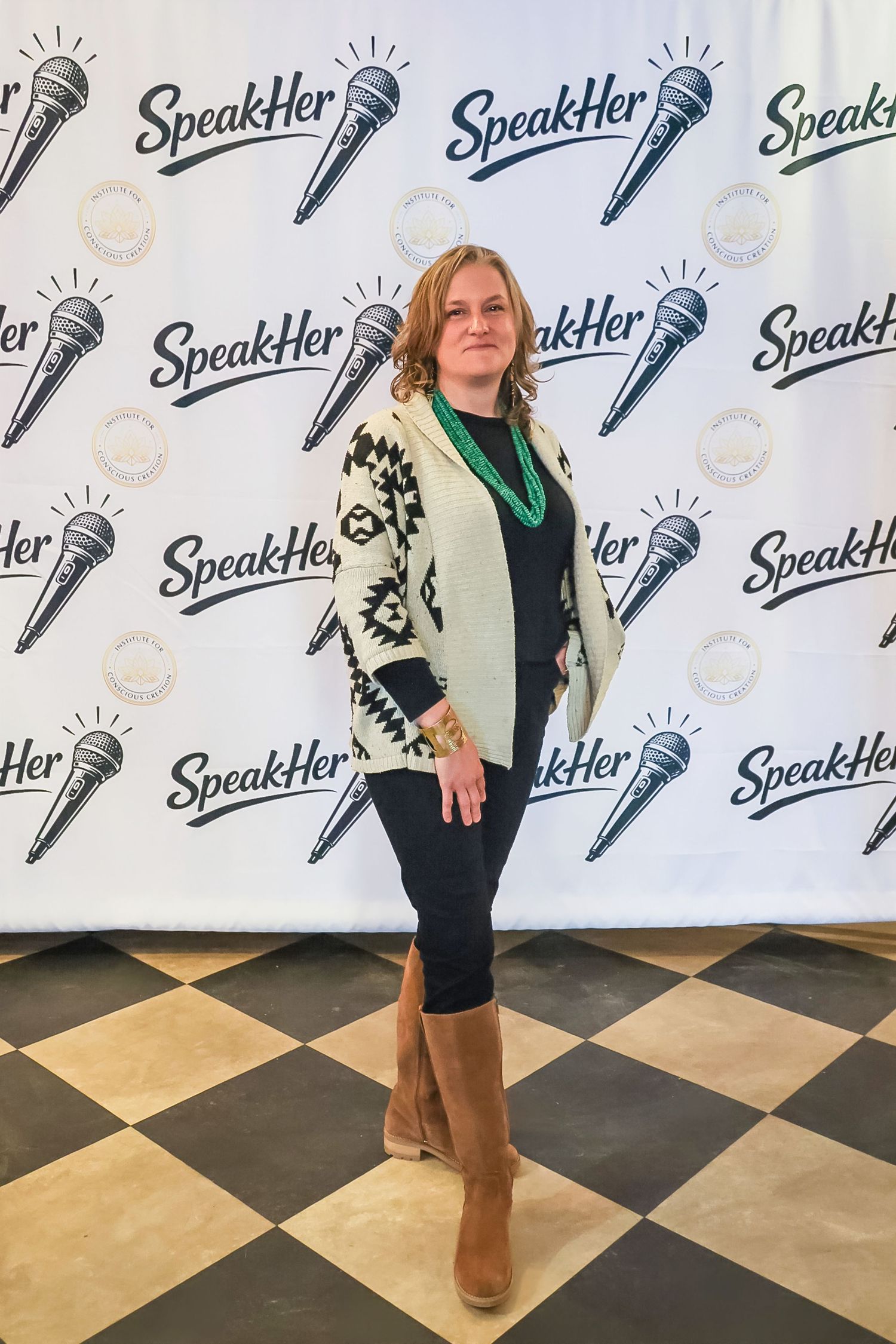 Woman in patterned cardigan and teal necklace posing at SpeakHer event backdrop on checkered floor.