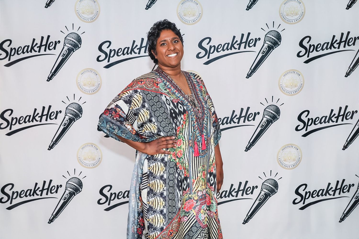 Woman in colorful patterned dress posing at SpeakHer event backdrop.
