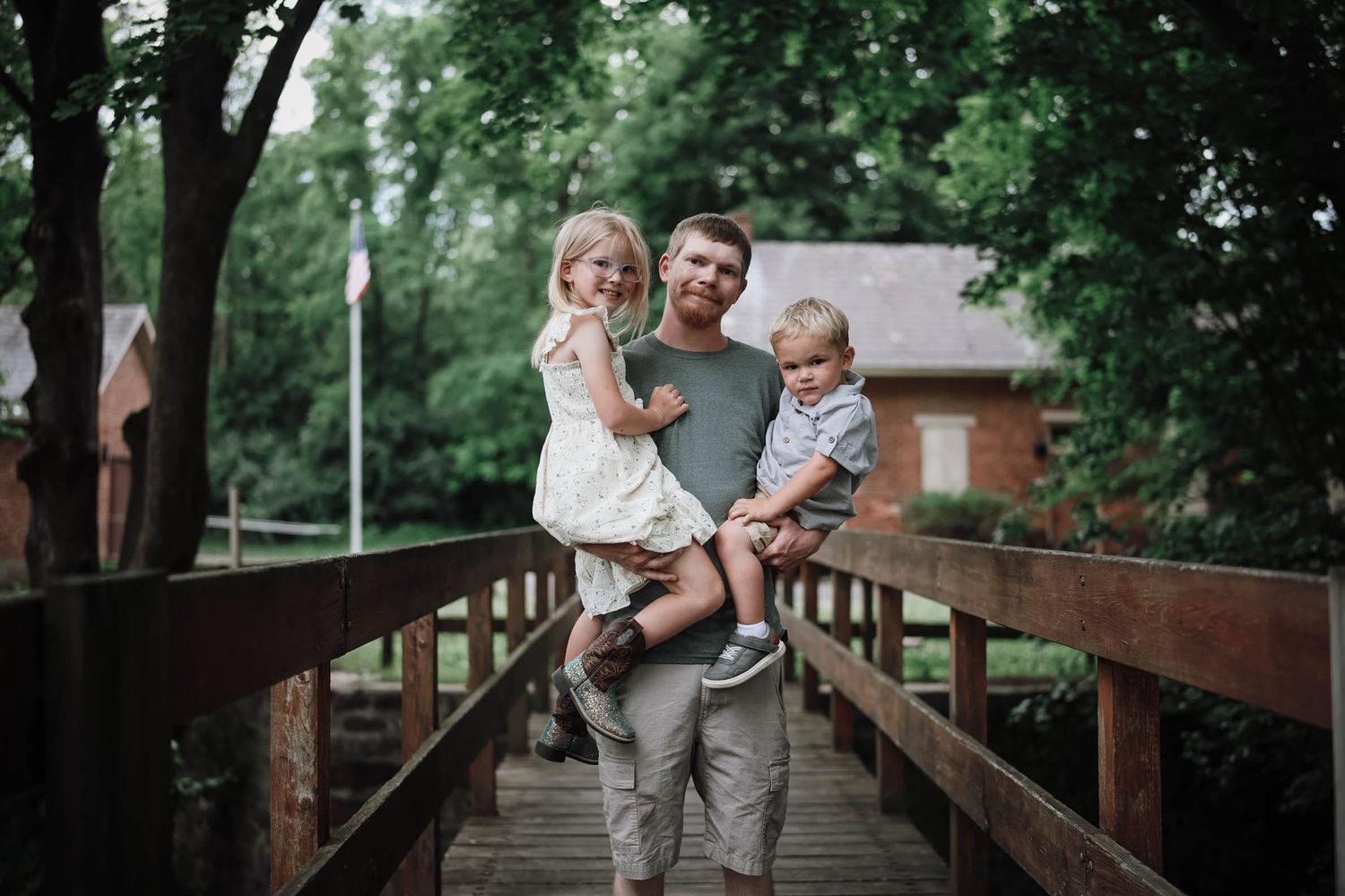 Father holding two young children on a wooden bridge surrounded by green trees.