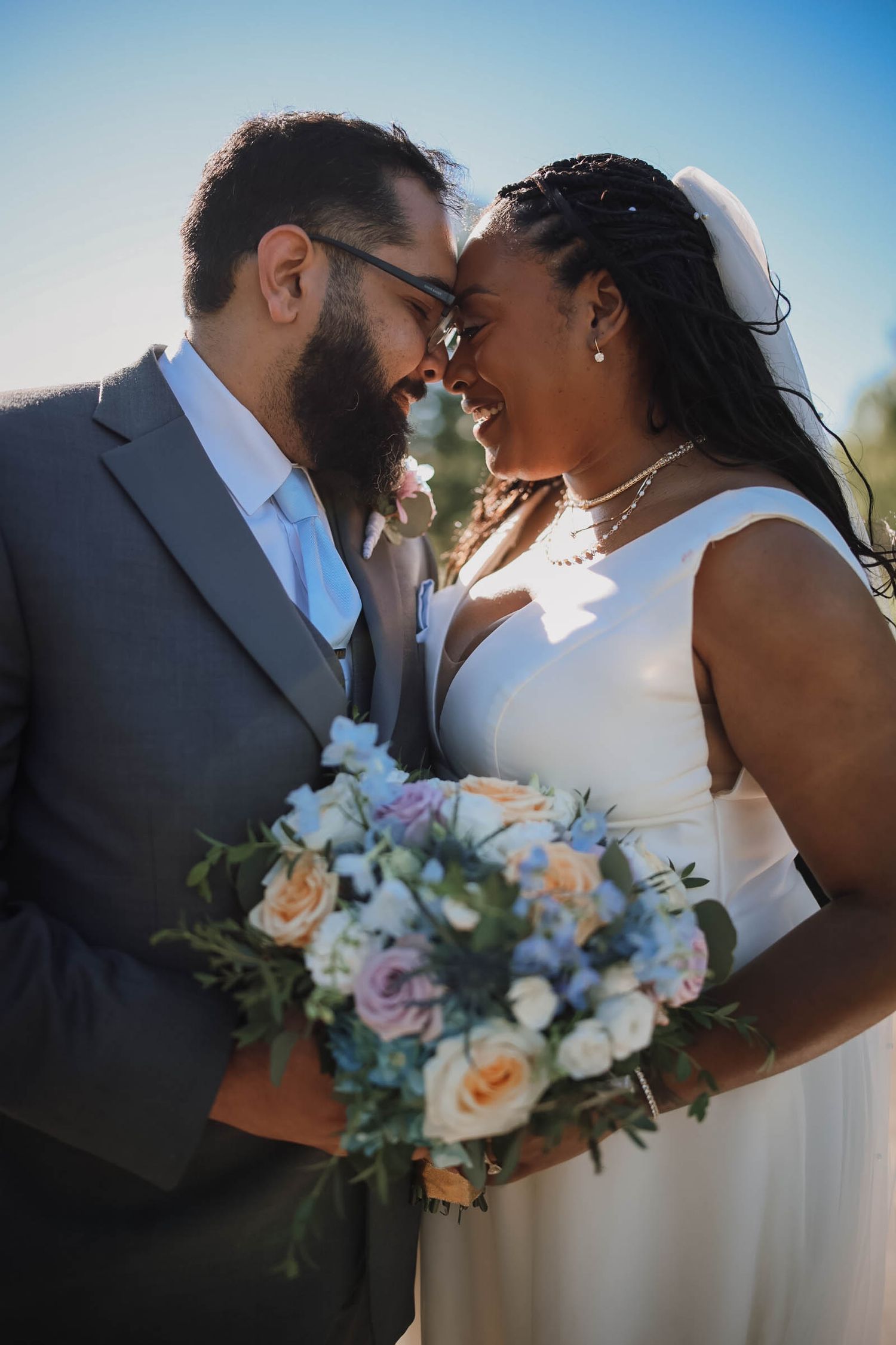 Interracial couple sharing a joyful moment on their wedding day, bride holding a pastel floral bouquet under a clear blue sky.
