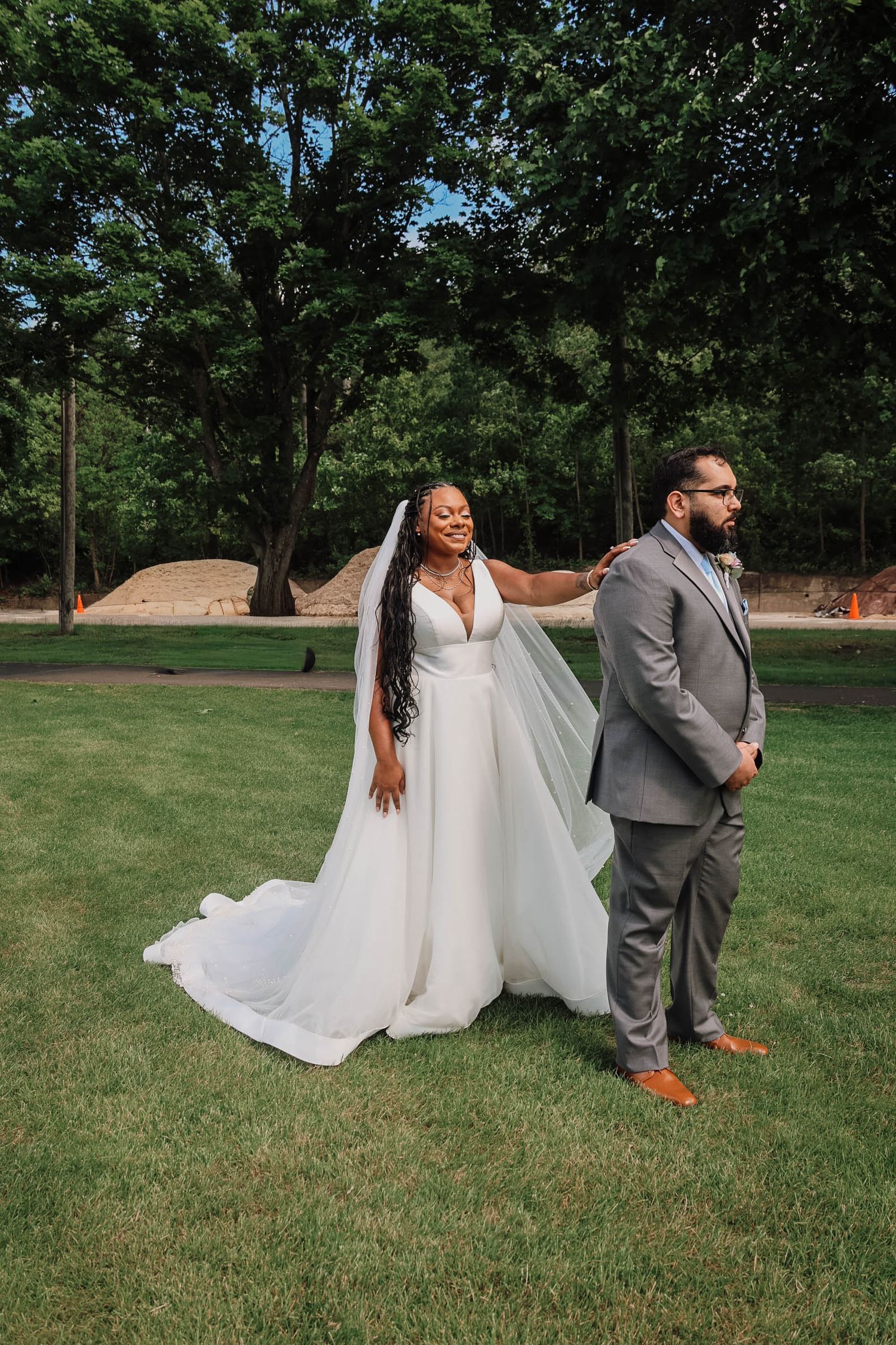 Bride in white gown and veil touches groom's shoulder in gray suit during outdoor first look on green lawn.
