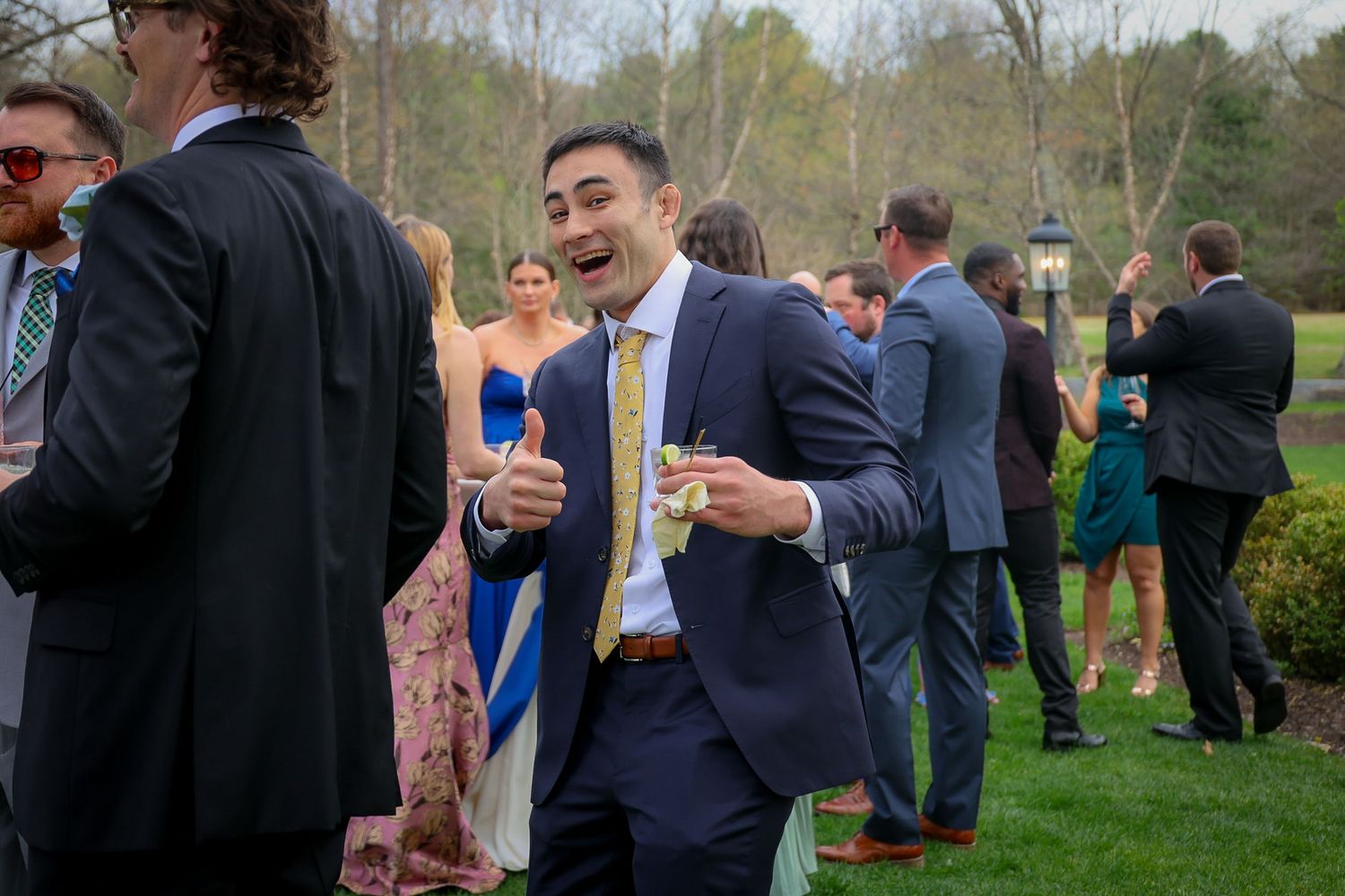 Man in navy suit with yellow tie laughing at outdoor wedding cocktail hour on green lawn.