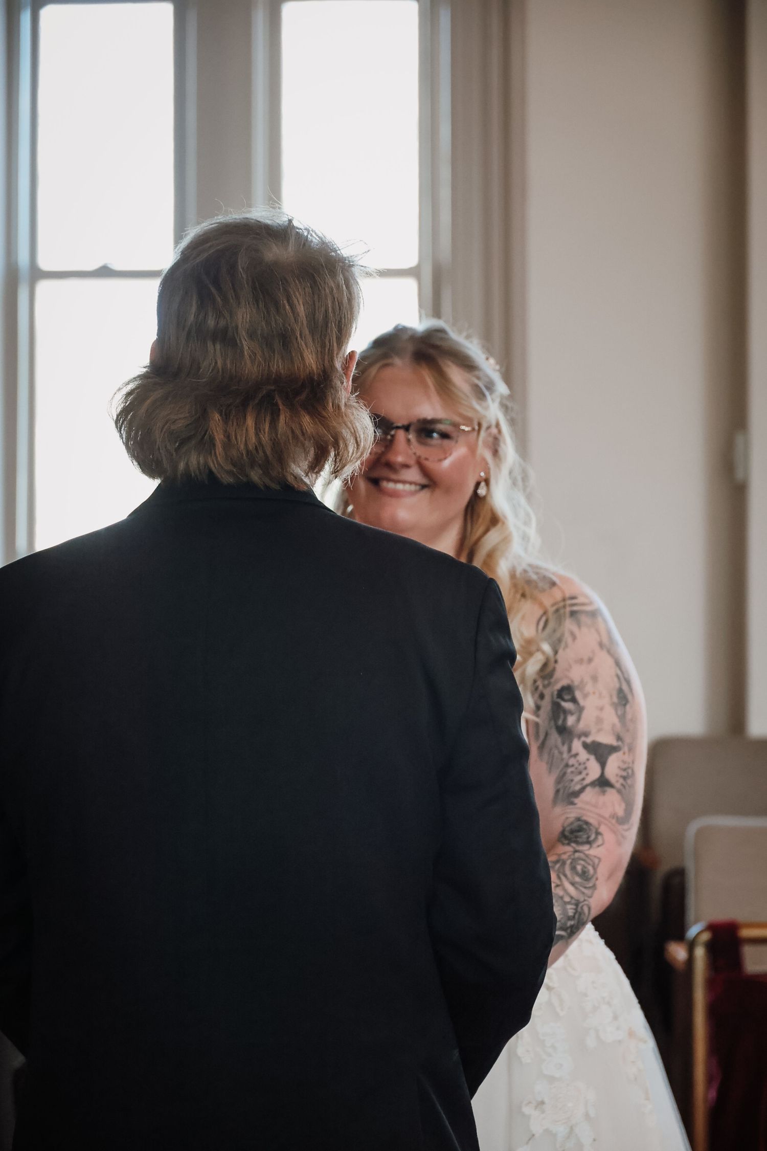 Bride with tattoos smiling at groom during indoor wedding ceremony near window.