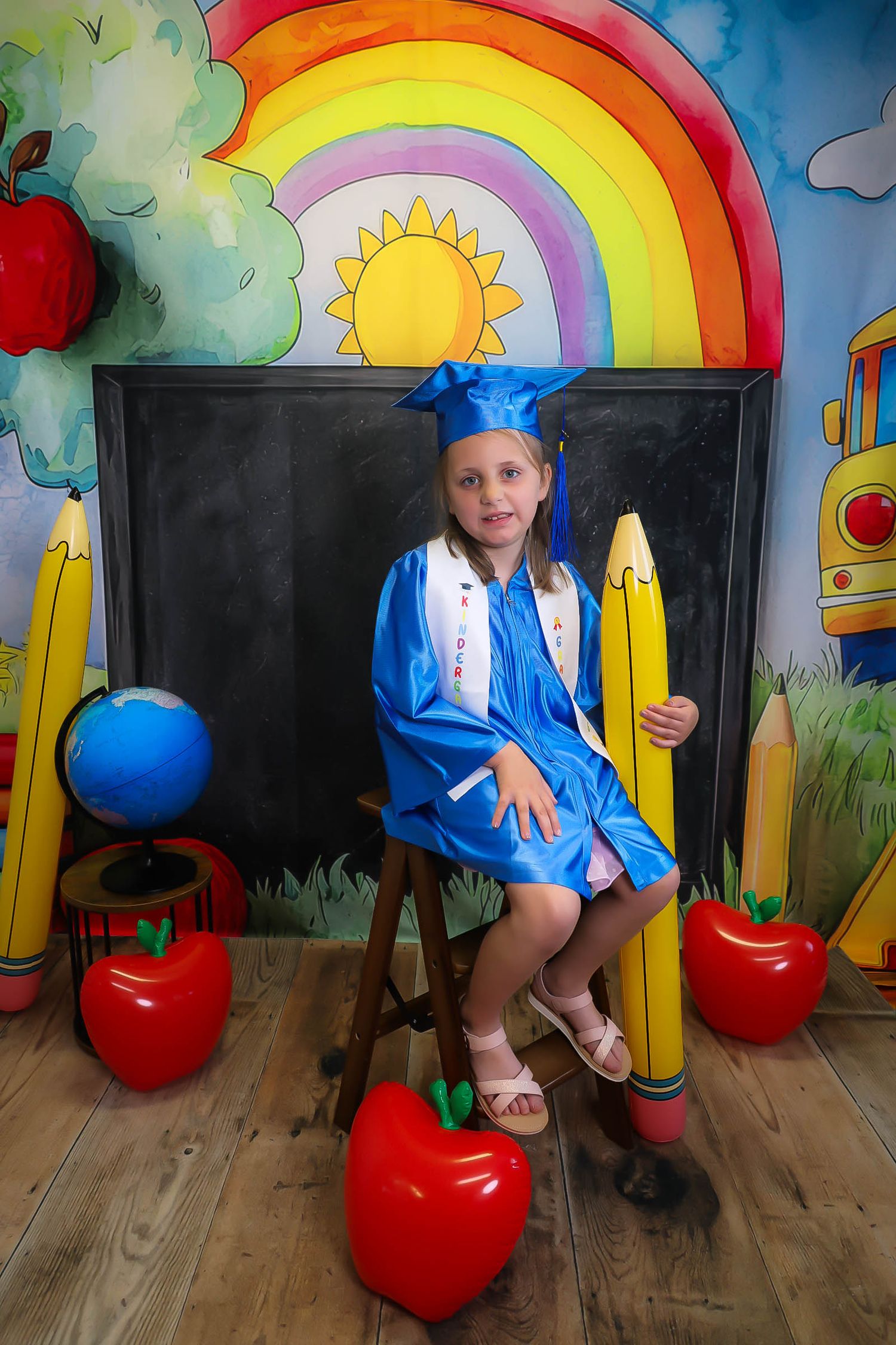 Young girl in blue graduation cap and gown sits proudly before colorful school-themed backdrop with pencils, apples, and rainbow.