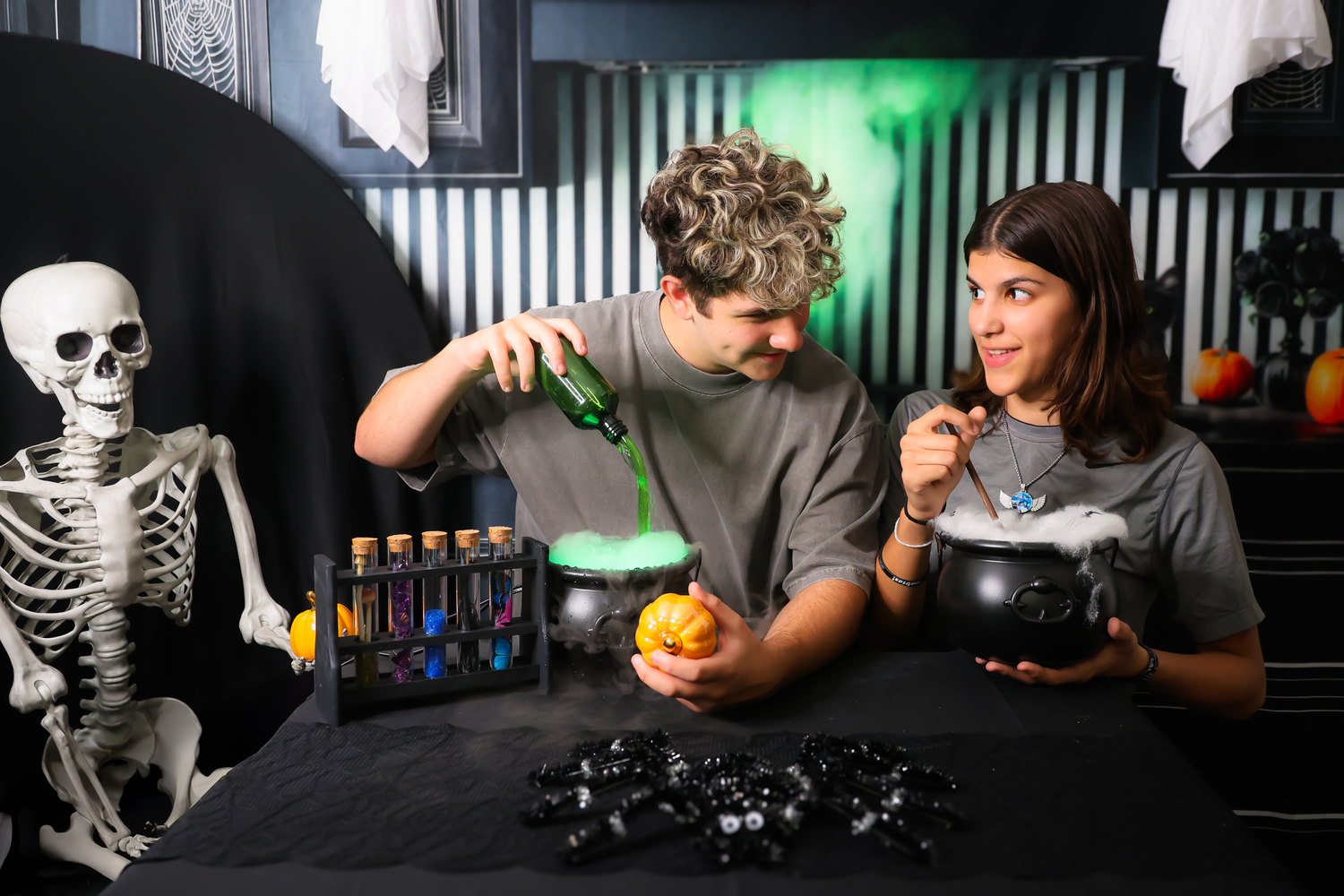 Two people doing Halloween science experiments with colorful potions, a cauldron, and skeleton decorations on a dark table.