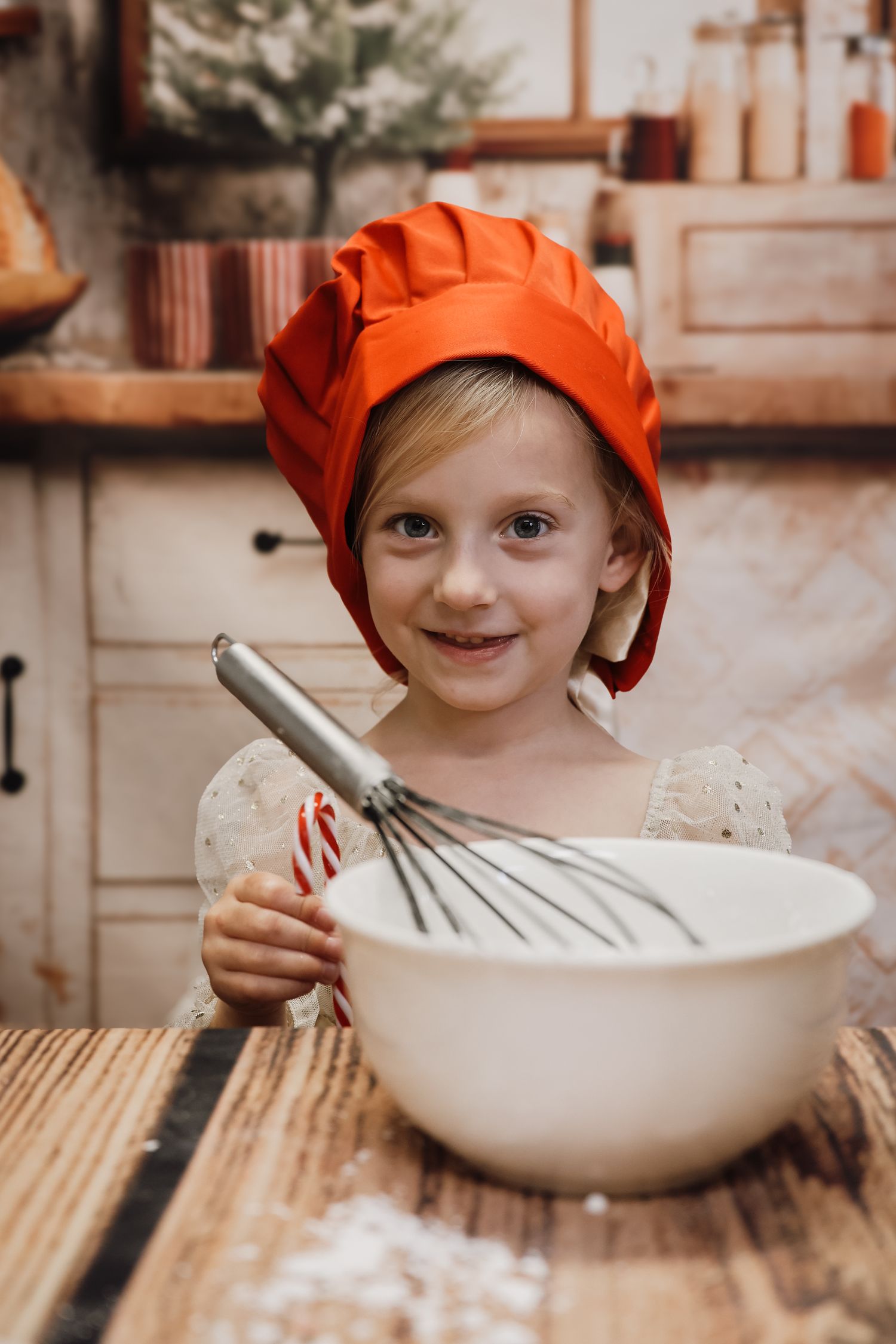 Young girl in red chef hat smiles while whisking batter in white bowl at wooden kitchen counter.