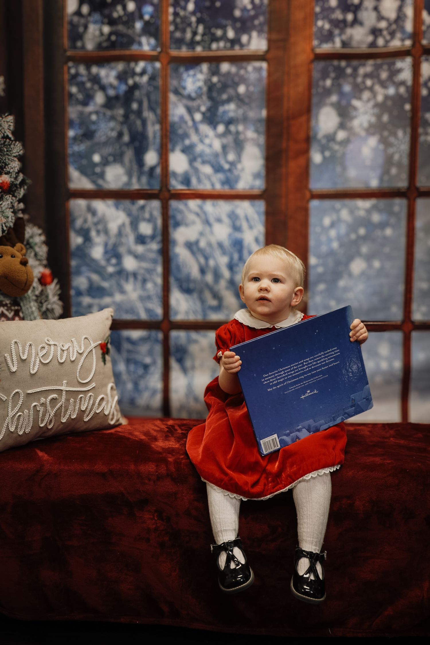 Baby girl in red dress holds a blue book on a festive red bench with a Merry Christmas pillow and snowy window backdrop.
