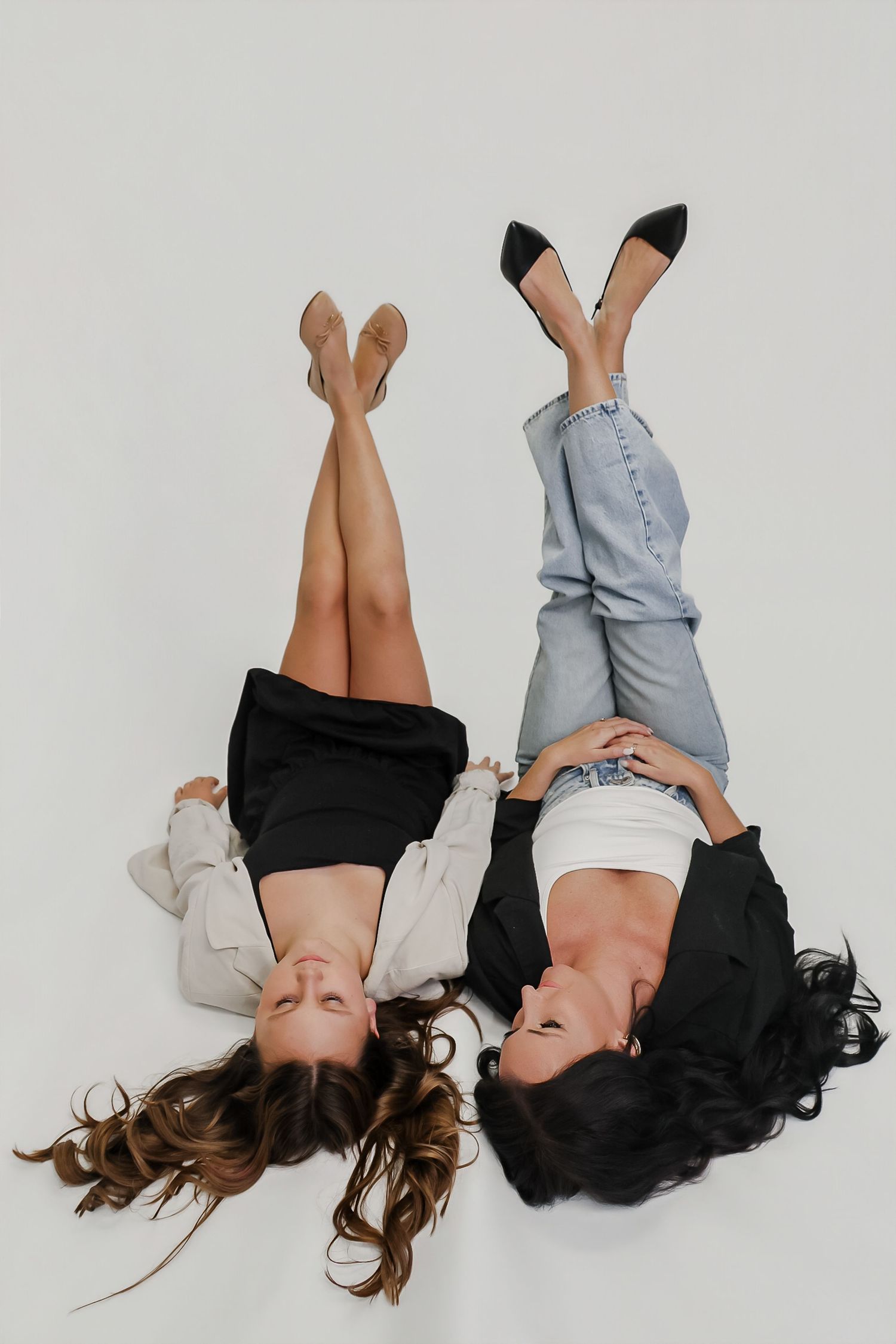 Two women lying on white floor with legs raised, one in jeans and one in black skirt, wearing heels.
