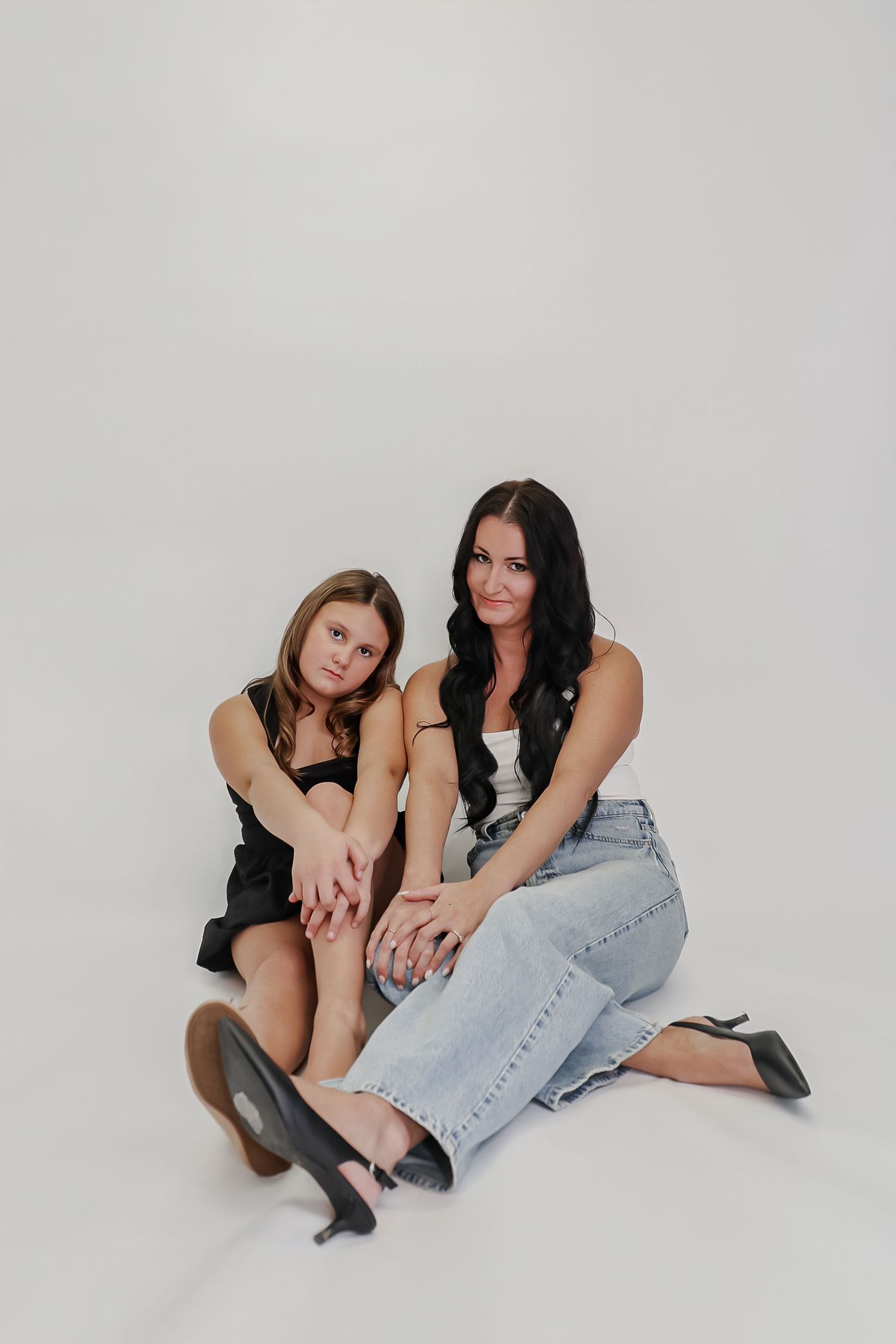 Two women sitting on white floor, one in black dress, one in jeans and white top, posing together for a studio photo.