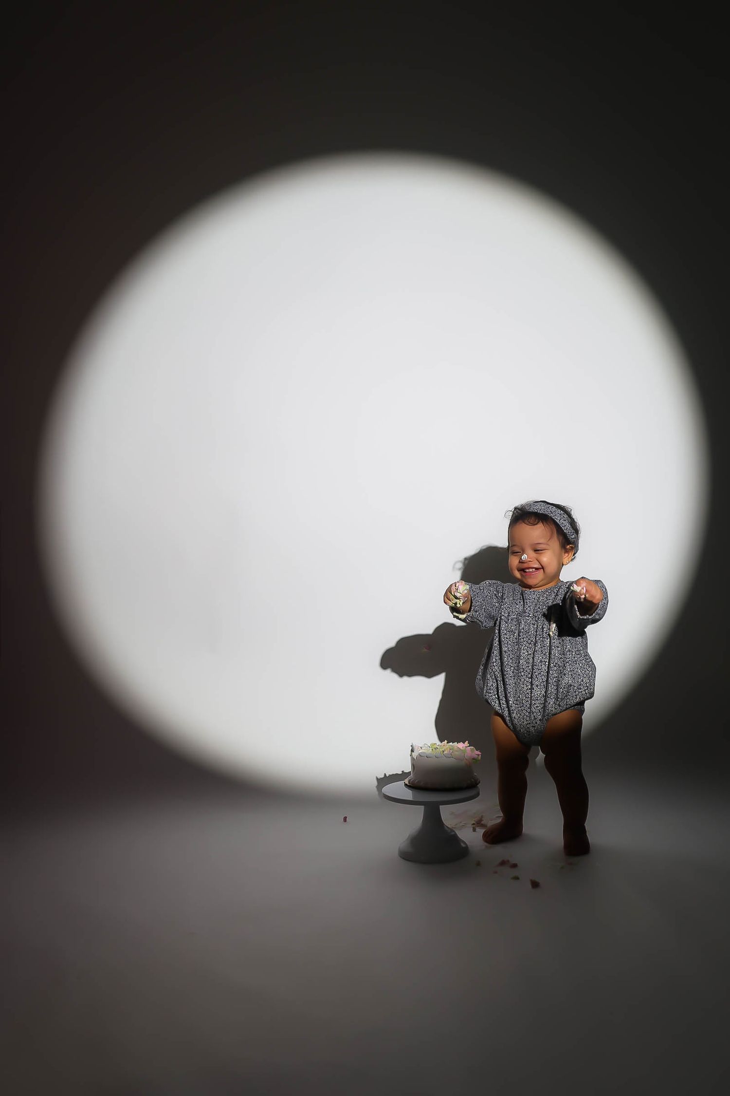 Toddler in gray romper celebrates first birthday smashing cake under dramatic spotlight.
