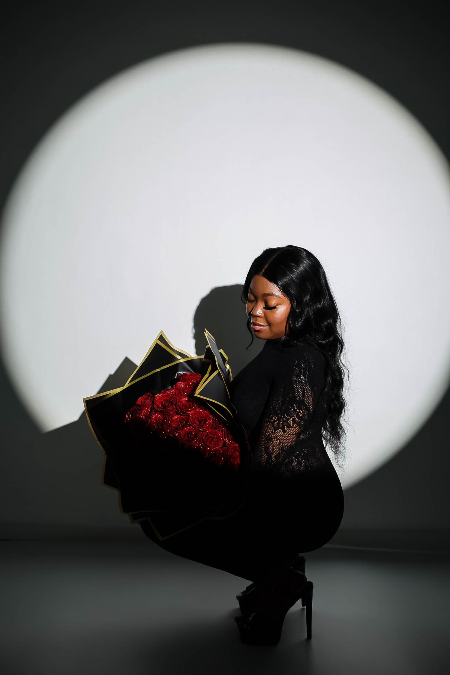 Black woman in lace outfit crouching while holding elegant wrapped red roses bouquet in studio spotlight.