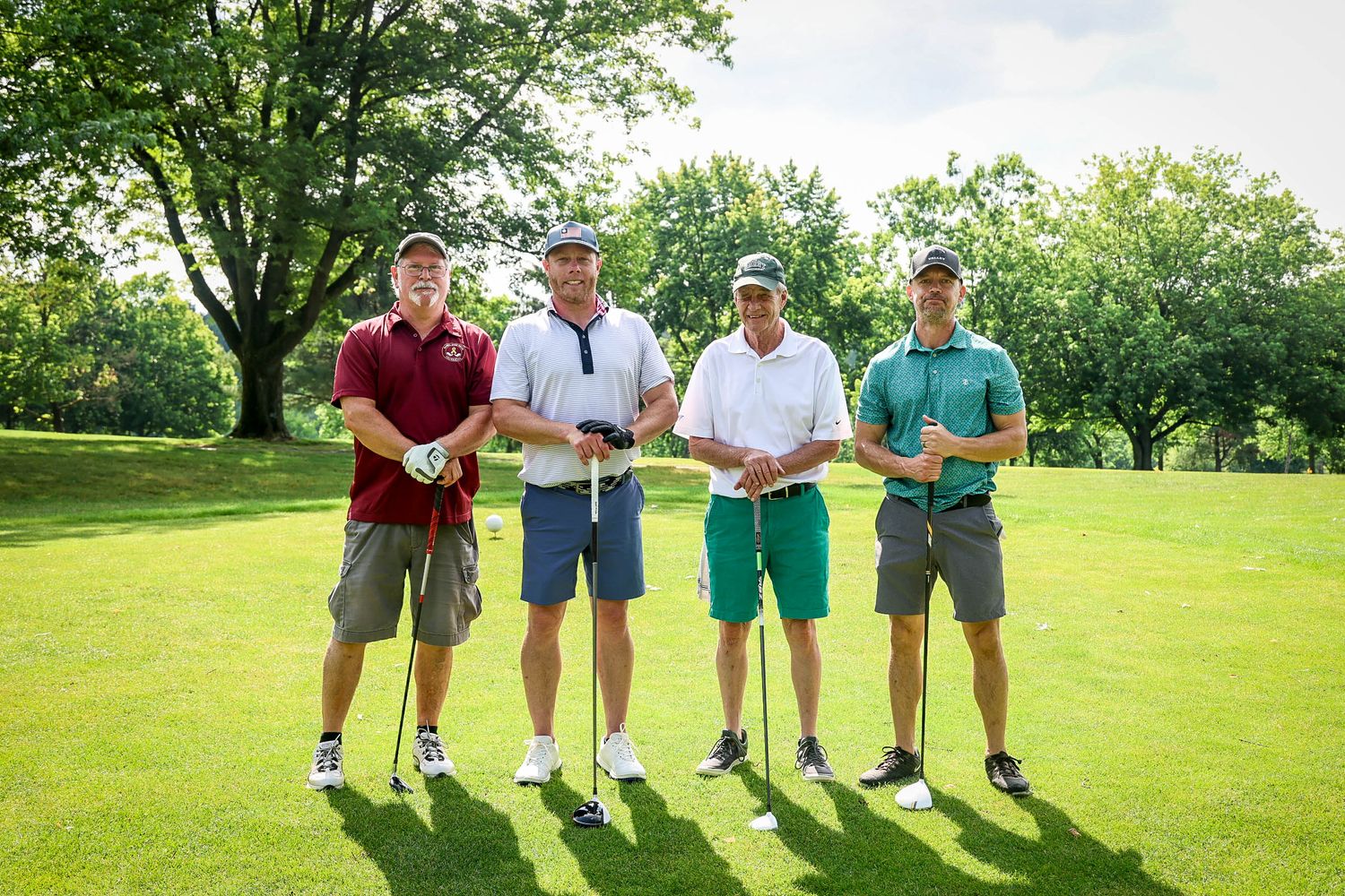 Four golfers pose together on a sunny green golf course, holding clubs and smiling for a group photo.