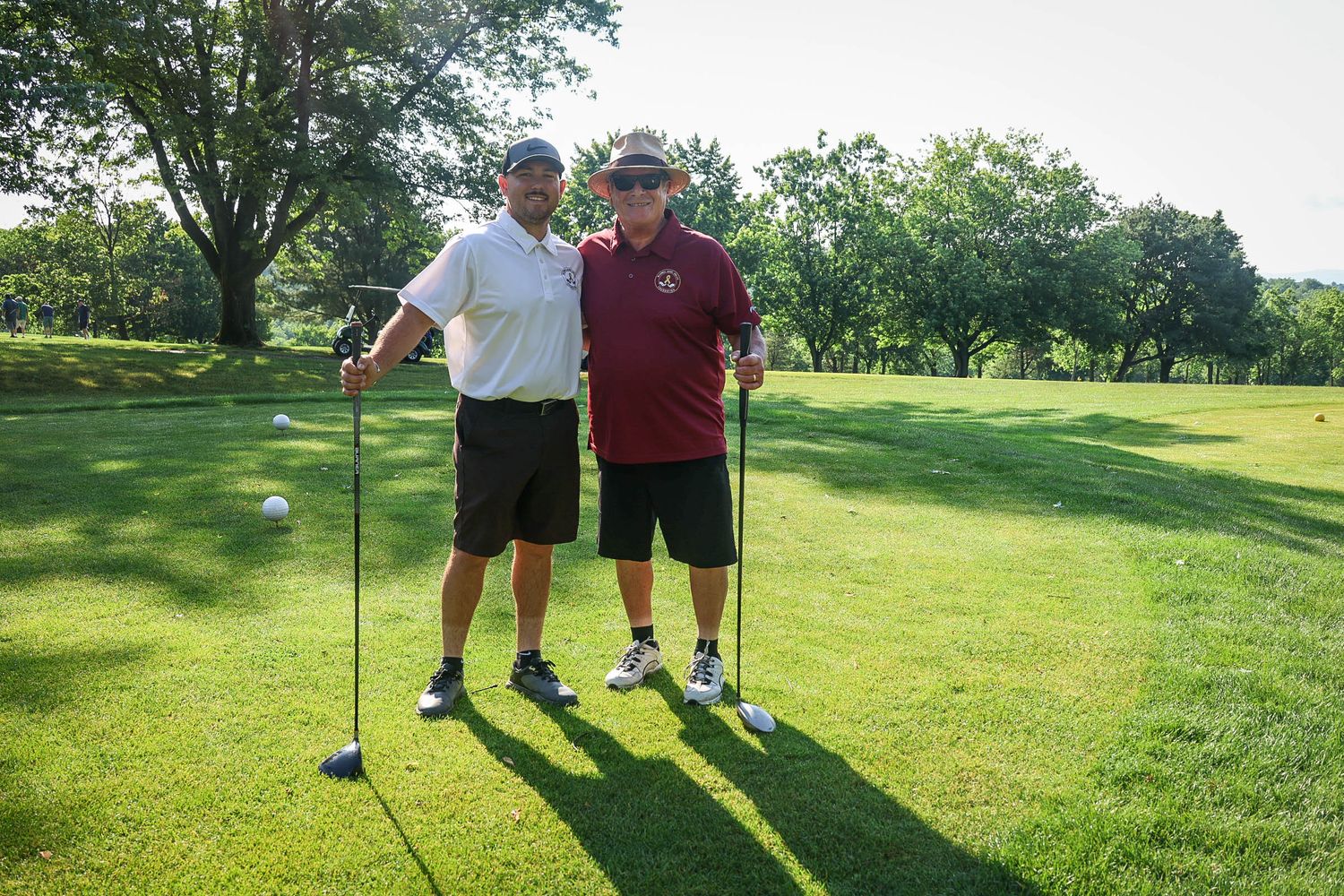 Two golfers posing with clubs on a sunny green golf course surrounded by trees.