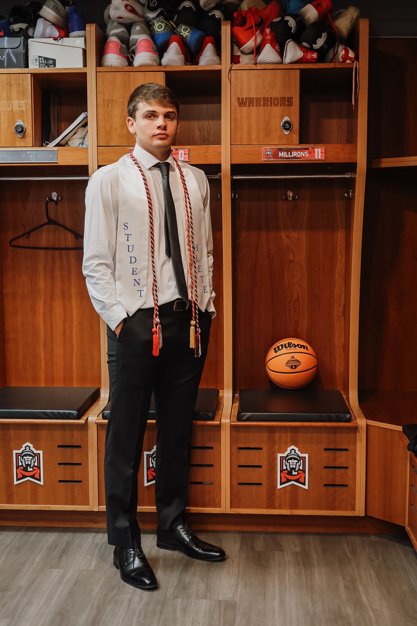 Young man in dress shirt and tie wearing student stole stands in basketball team locker room with Wilson ball.