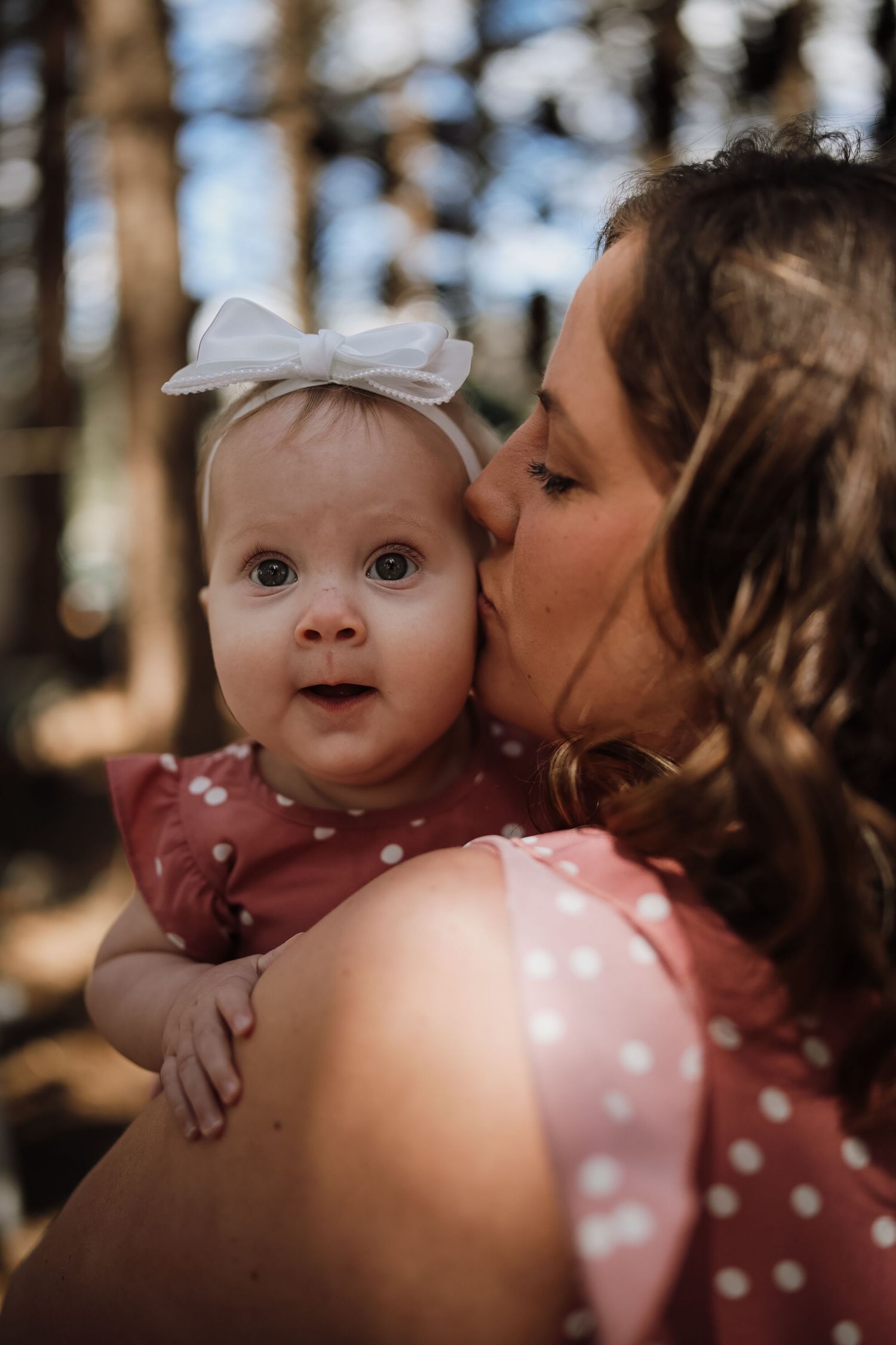 A mother kissing her baby girl wearing a white bow headband and pink polka dot dress in a forest setting.