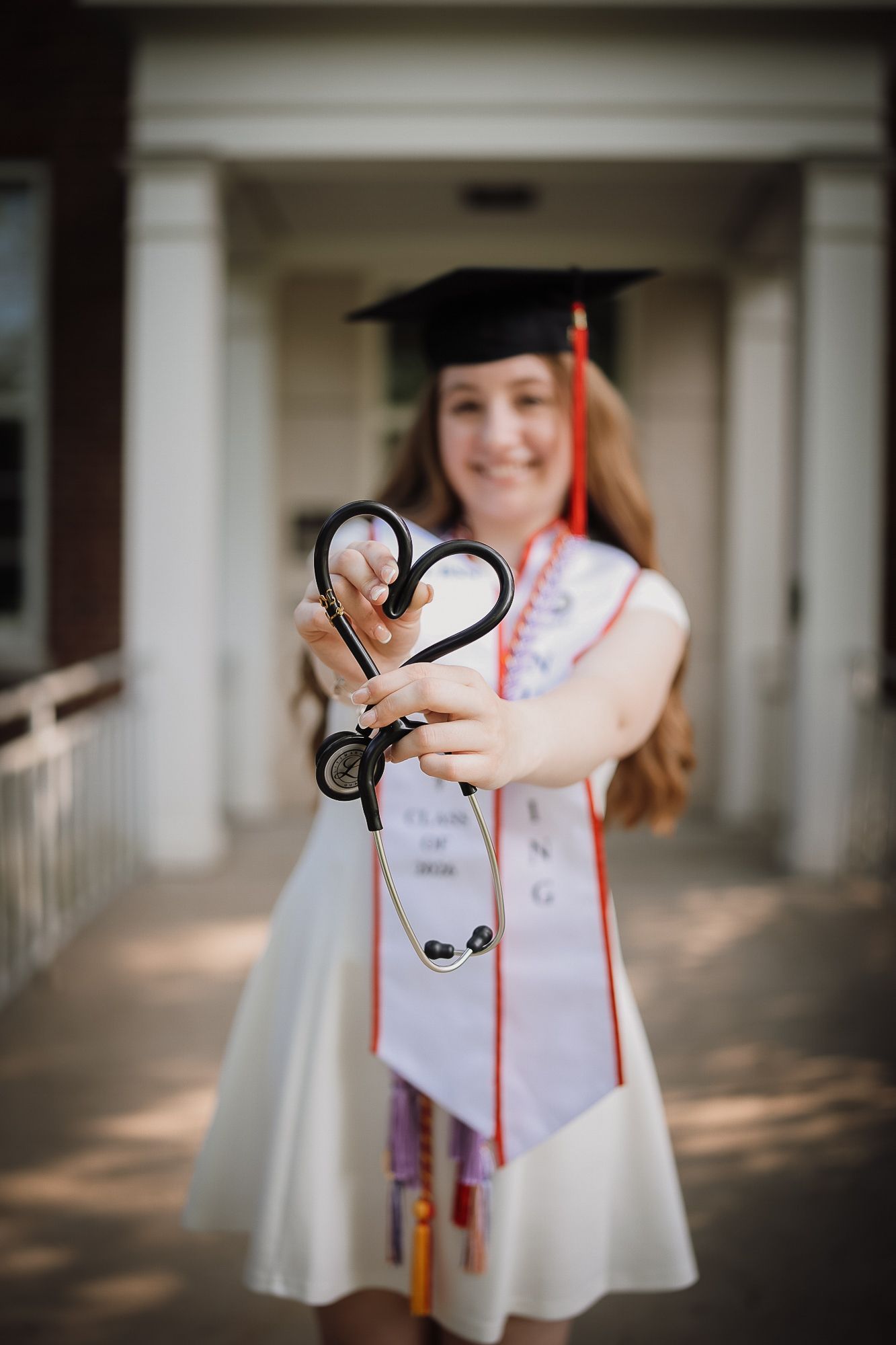 Nursing graduate in cap and gown holds stethoscope in heart shape toward camera at college building.