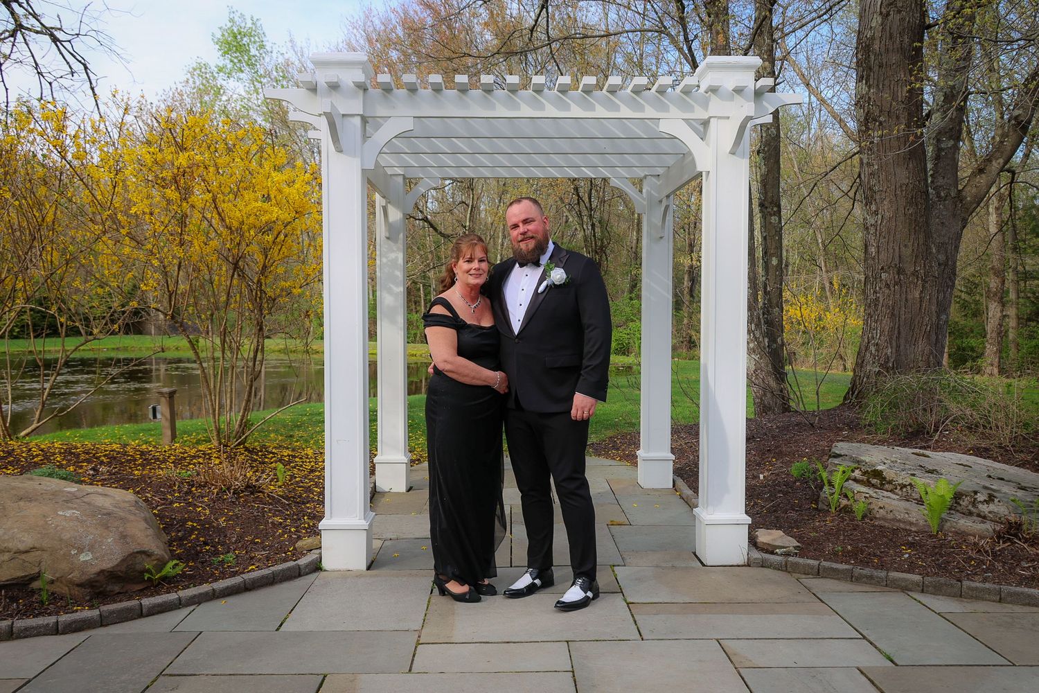 Couple in formal attire posing under white pergola in garden with spring foliage.