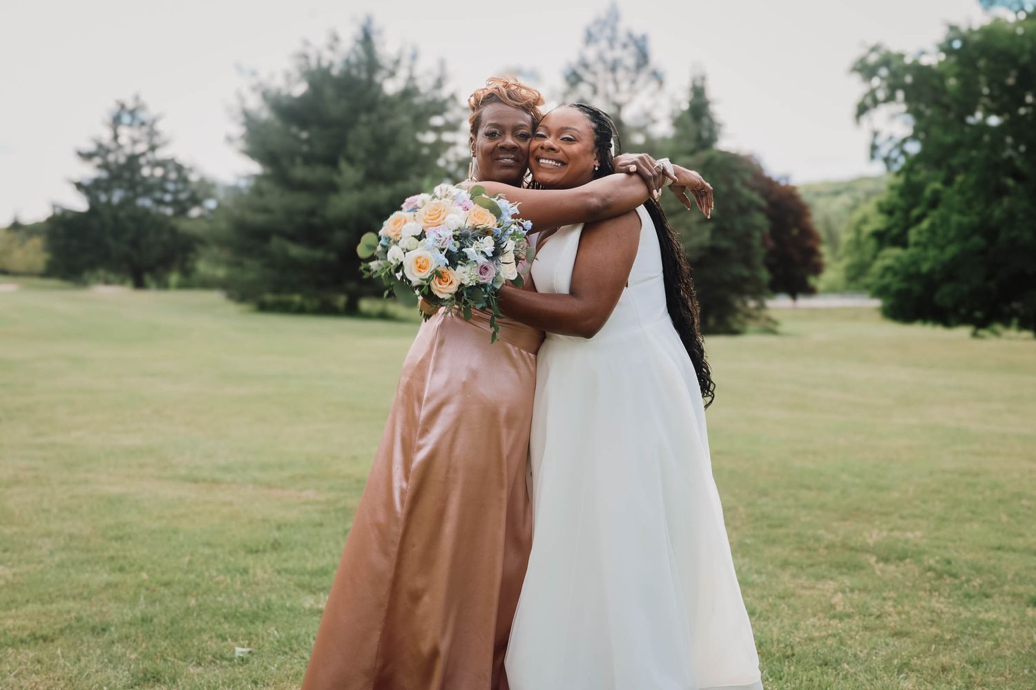 Two women in elegant dresses embrace joyfully outdoors, one holding a bouquet, celebrating a wedding day.
