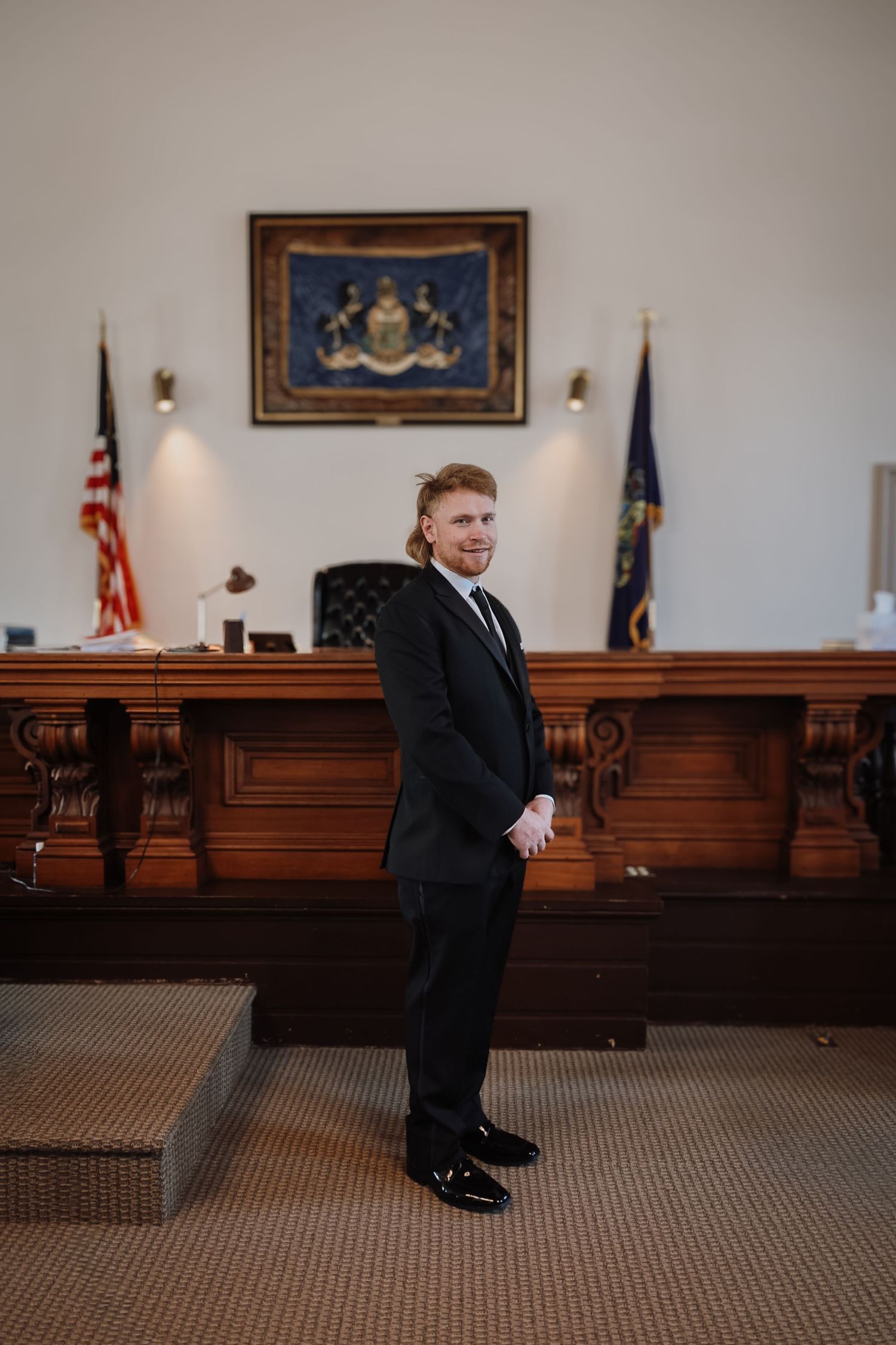 Groom in black tuxedo stands in elegant courtroom with wooden judge bench, American flag, and state seal painting.