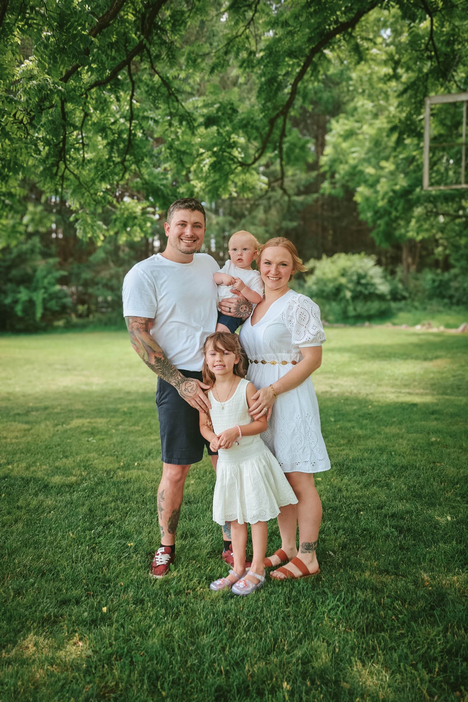 Happy family of four posing outdoors on green grass under trees, parents holding young children in white summer outfits.