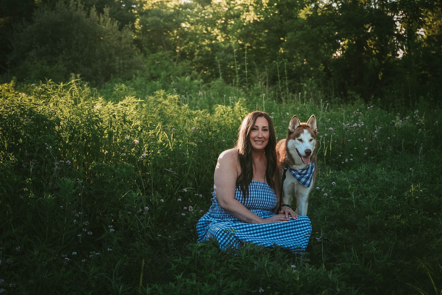 Woman in blue gingham dress smiling with Siberian Husky dog in lush green meadow at golden hour.