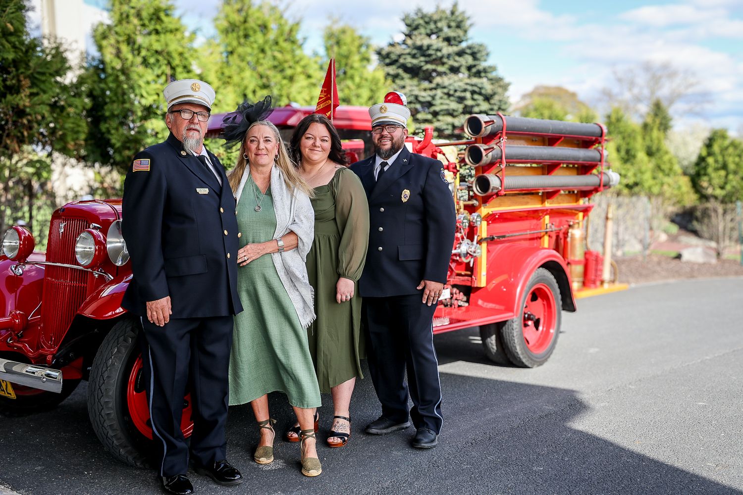 Four people pose beside a vintage red fire engine, two in firefighter uniforms and two women in elegant dresses.