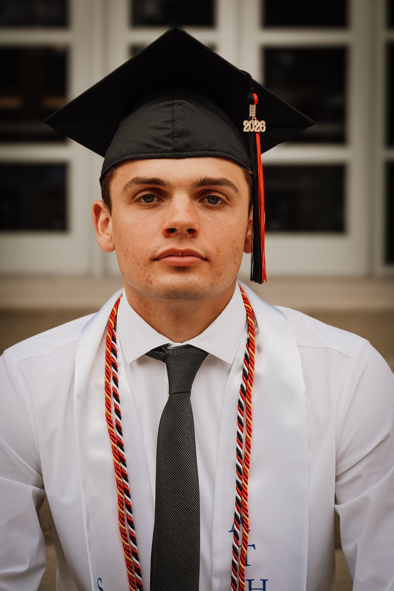 Young man in graduation cap and gown with honor cords poses for a 2026 graduation portrait photo.