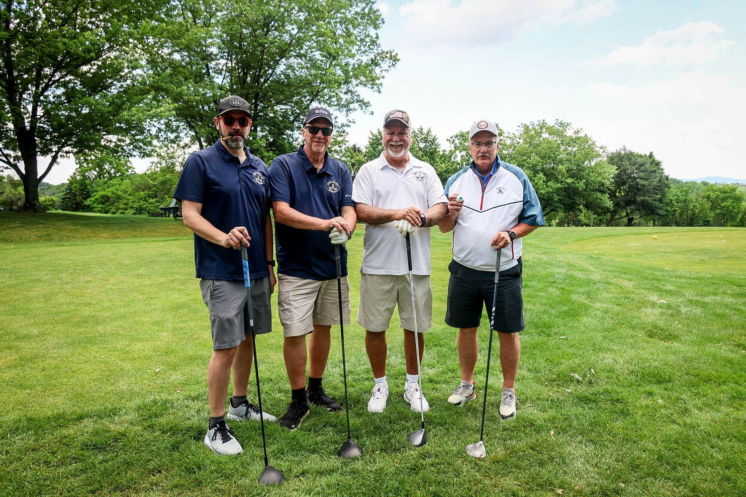 Four men posing with golf clubs on a lush green golf course on a sunny summer day.
