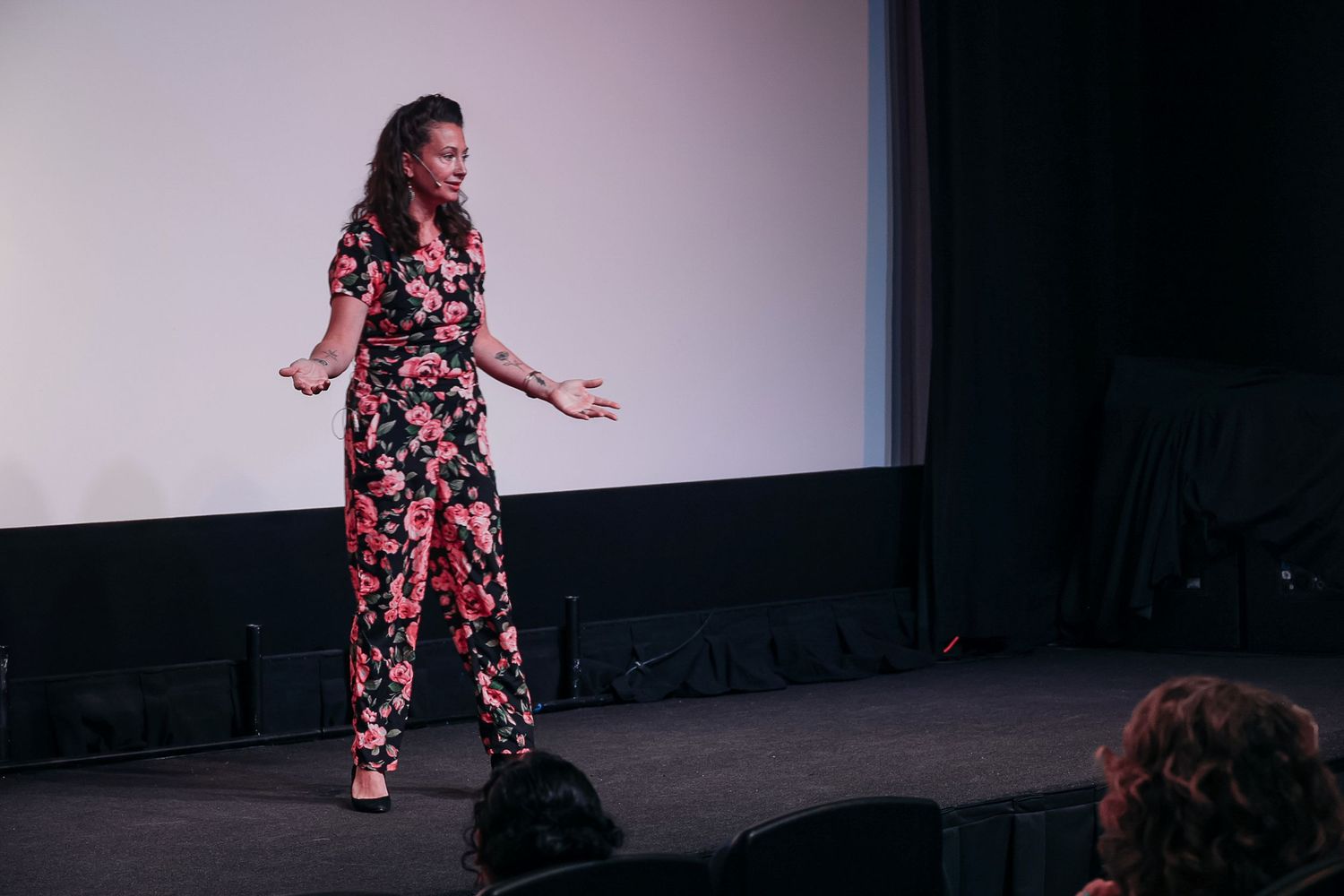 Woman in floral jumpsuit presenting on stage before a projection screen at an indoor event.