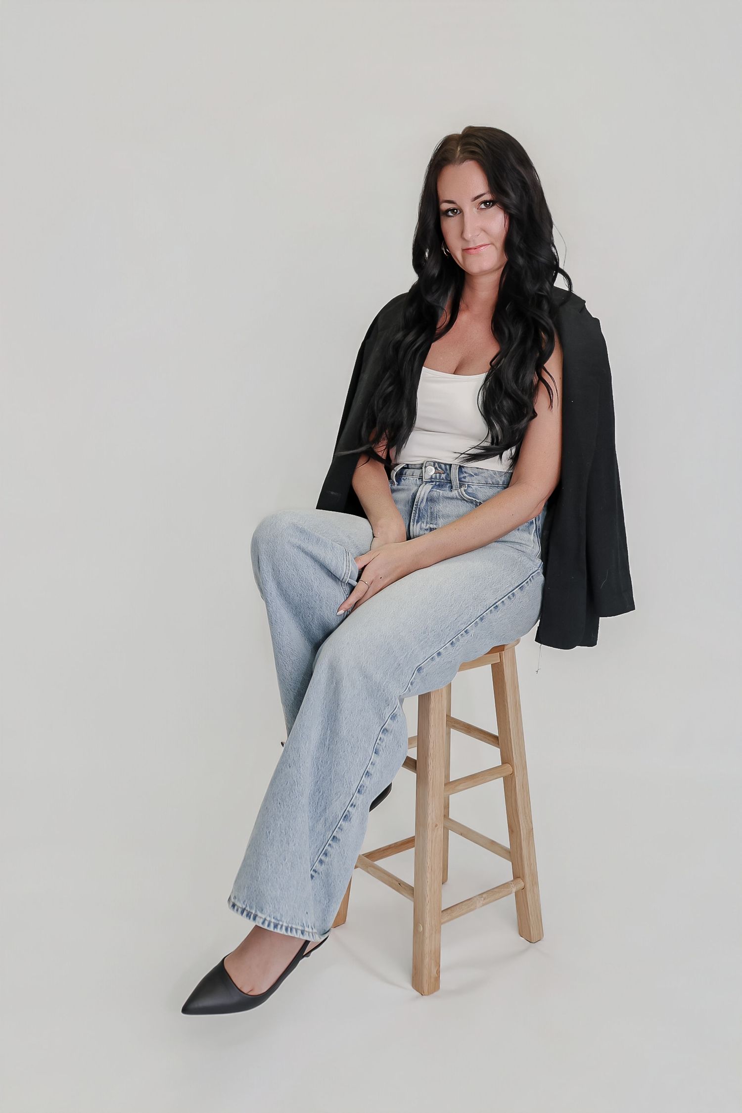 Woman with dark wavy hair sits on wooden stool wearing white top, light jeans, and black blazer draped over shoulders.