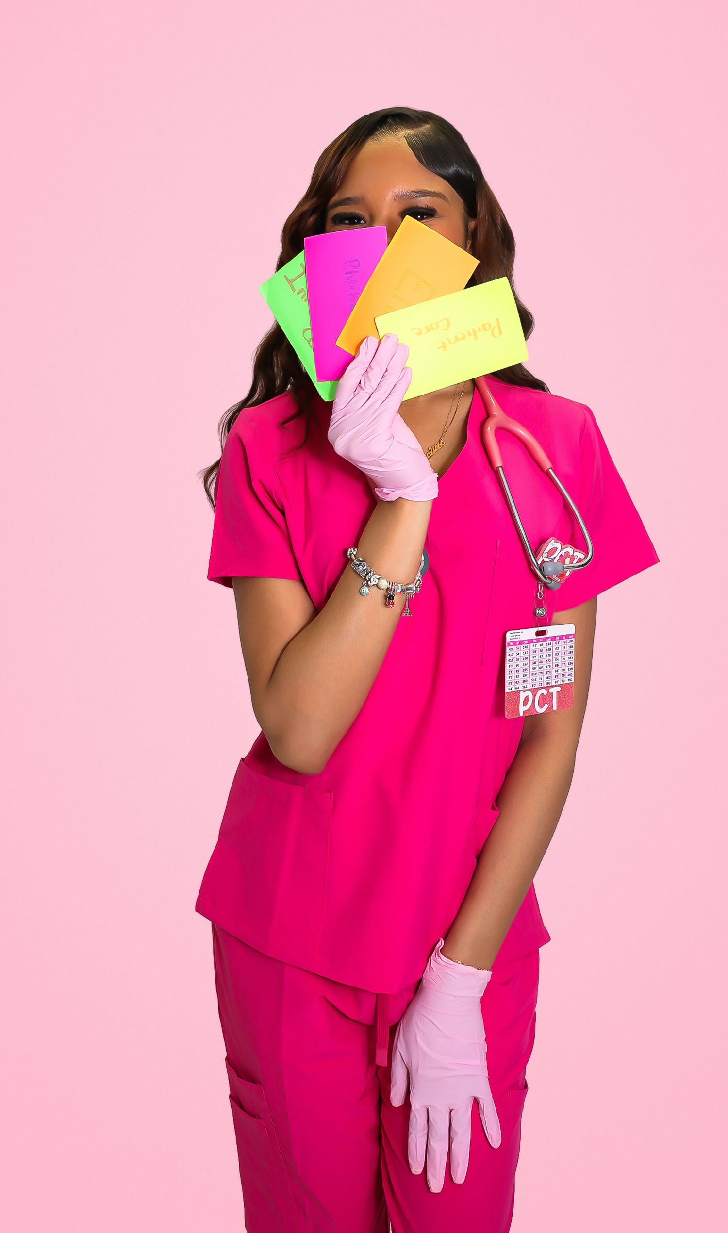 A female PCT nurse in pink scrubs and gloves holds colorful sticky notes while wearing a stethoscope against a pink background.