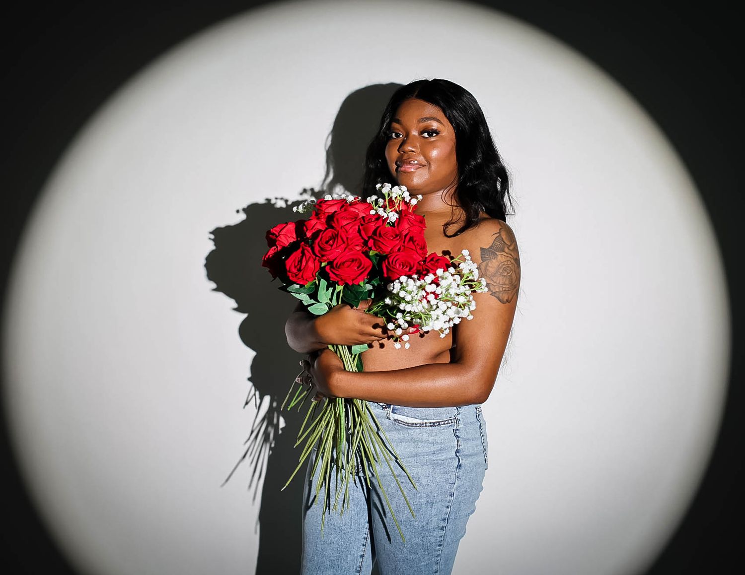 Young Black woman smiling, holding red roses bouquet, wearing jeans, illuminated by spotlight on white background.